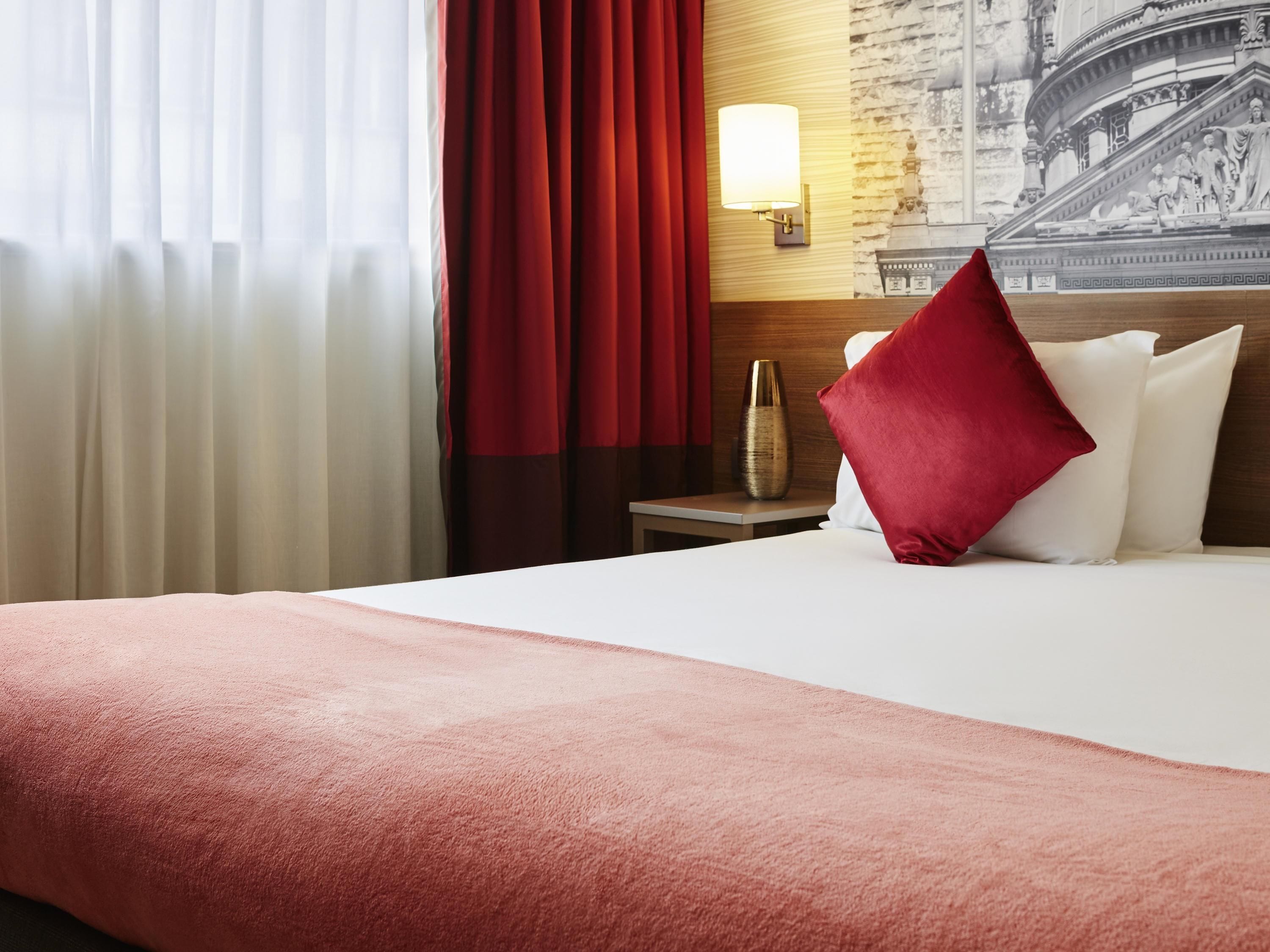 Een hotelkamer met een bed, rode gordijnen, een nachtkastje met een lamp en een wanddecoratie.