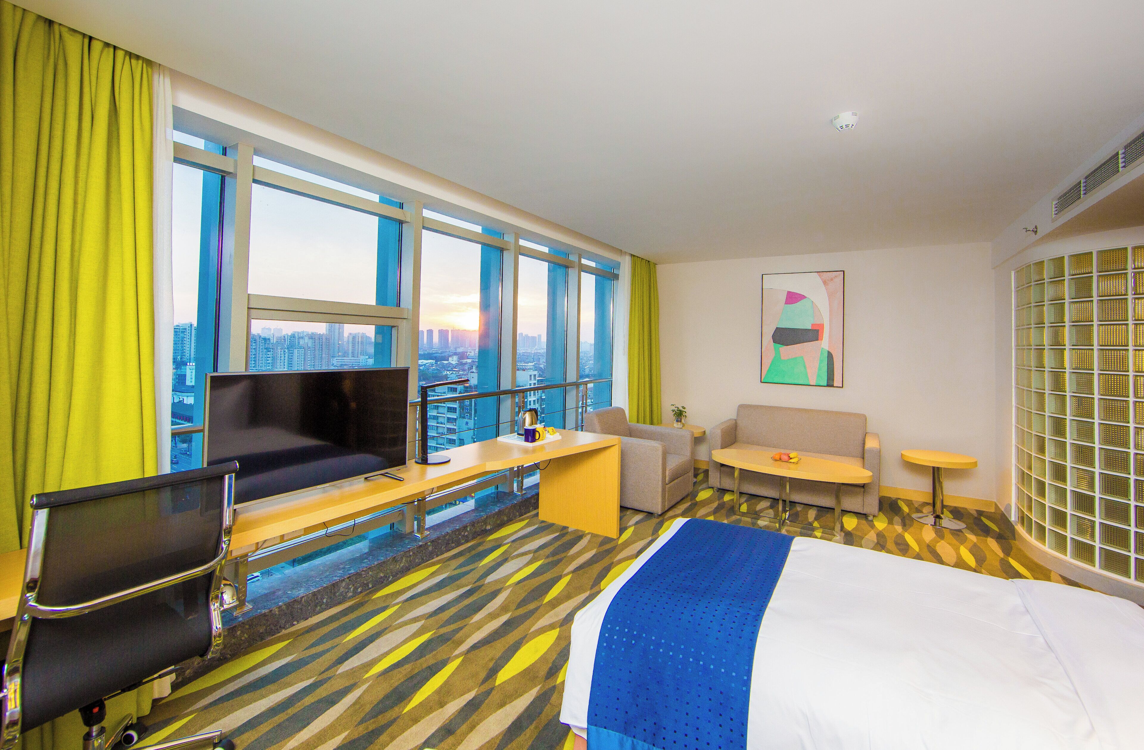 Habitación de hotel con cama, escritorio, silla, sofá, televisor y ventana grande con vista a la ciudad.