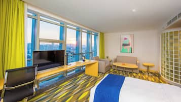 Habitación de hotel con cama, escritorio, silla, sofá, televisor y ventana grande con vista a la ciudad.