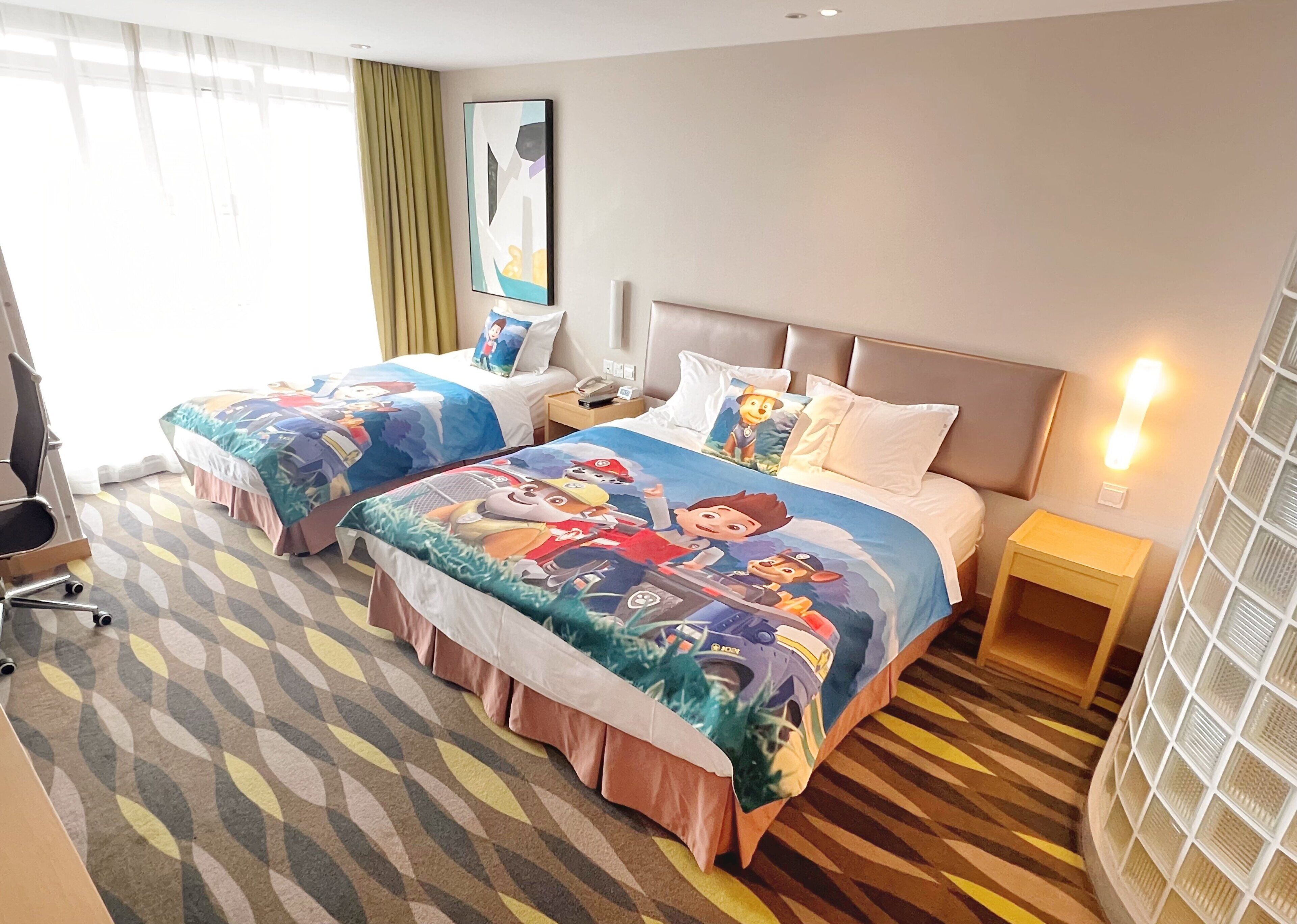 A hotel room with two beds, a desk, a chair, and a lamp.