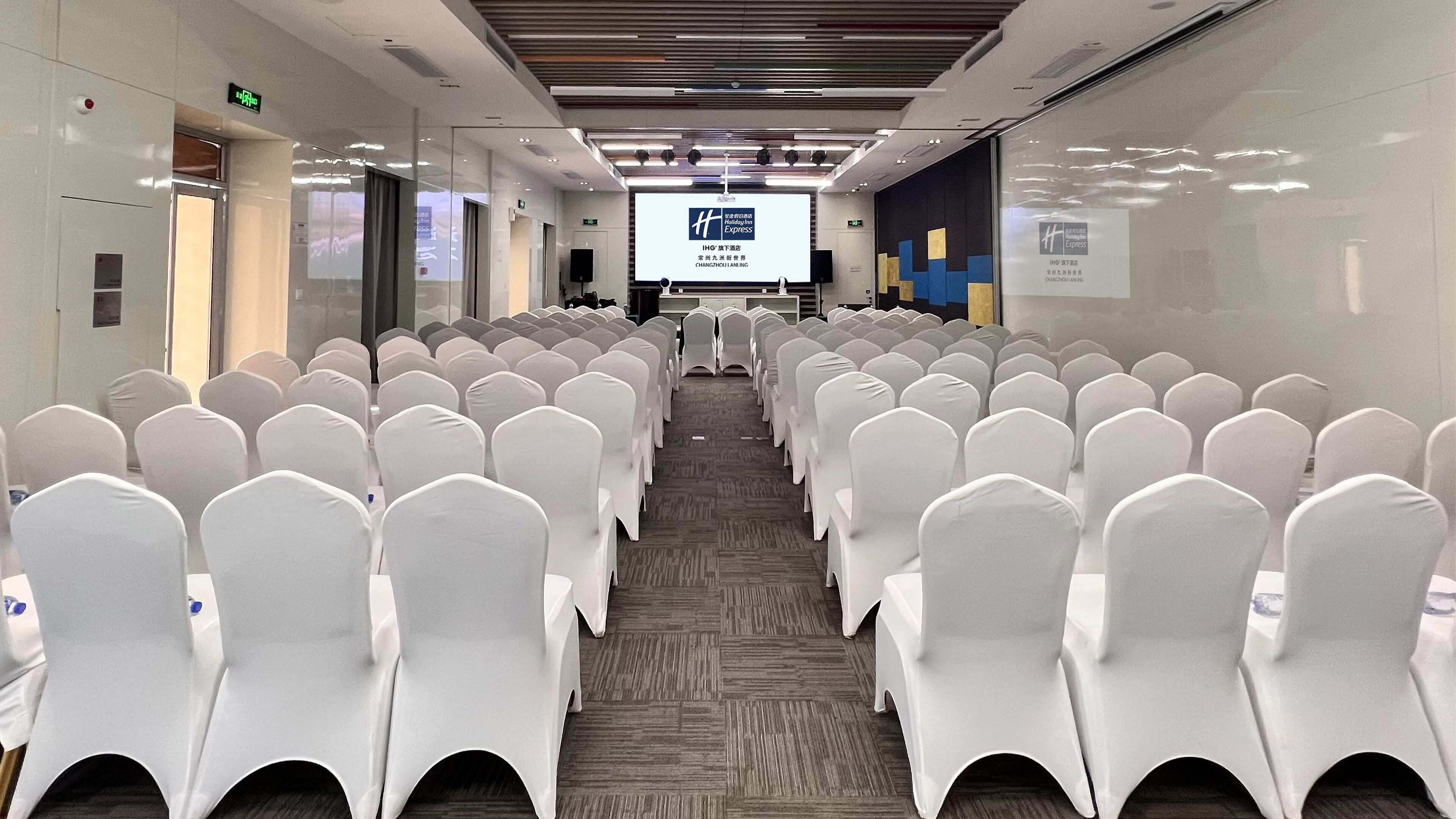 A conference room with rows of white chairs, a projection screen at the front, and a carpeted floor.