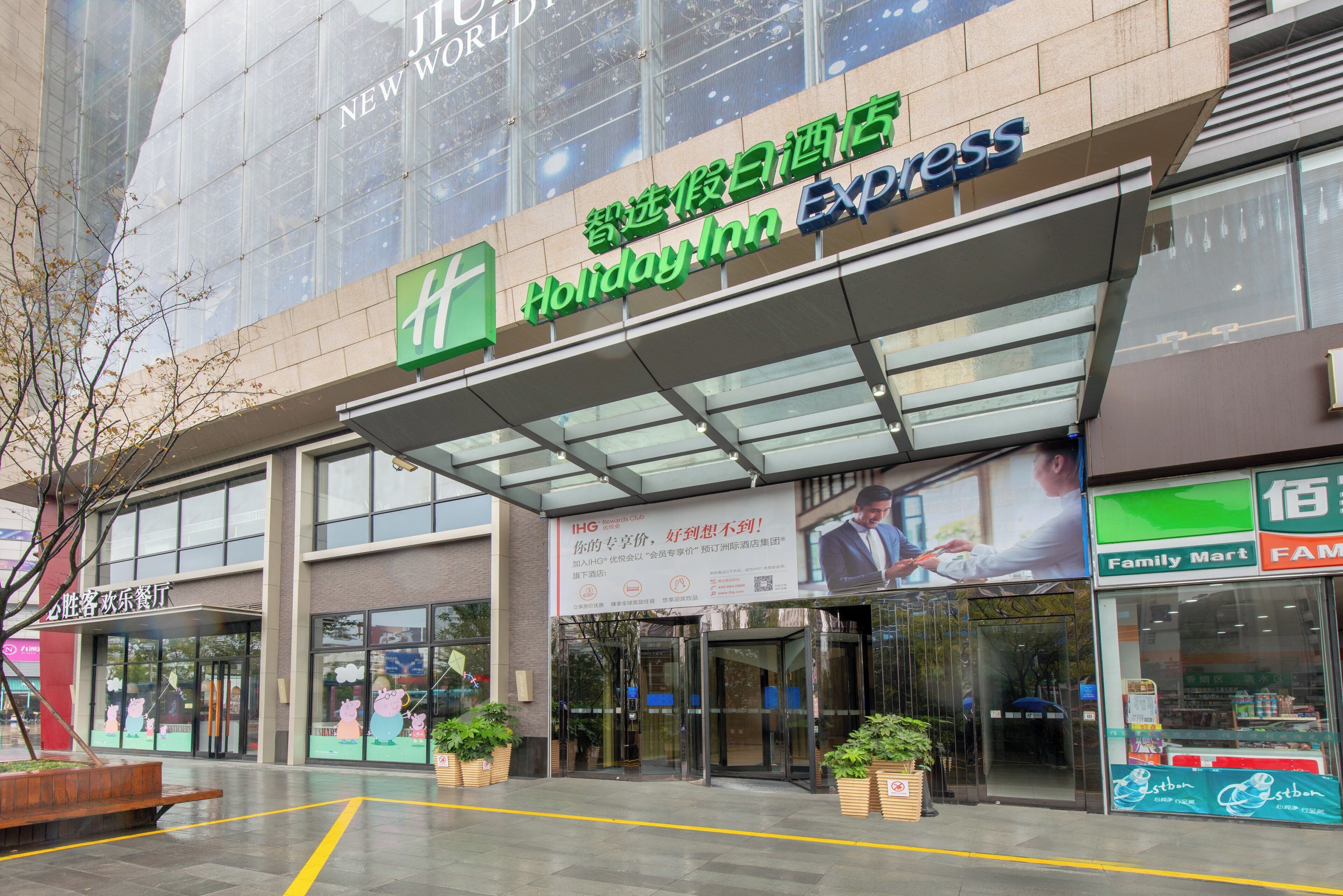 Un hotel Holiday Inn Express con fachada de vidrio, zona de estar al aire libre y entrada visible.