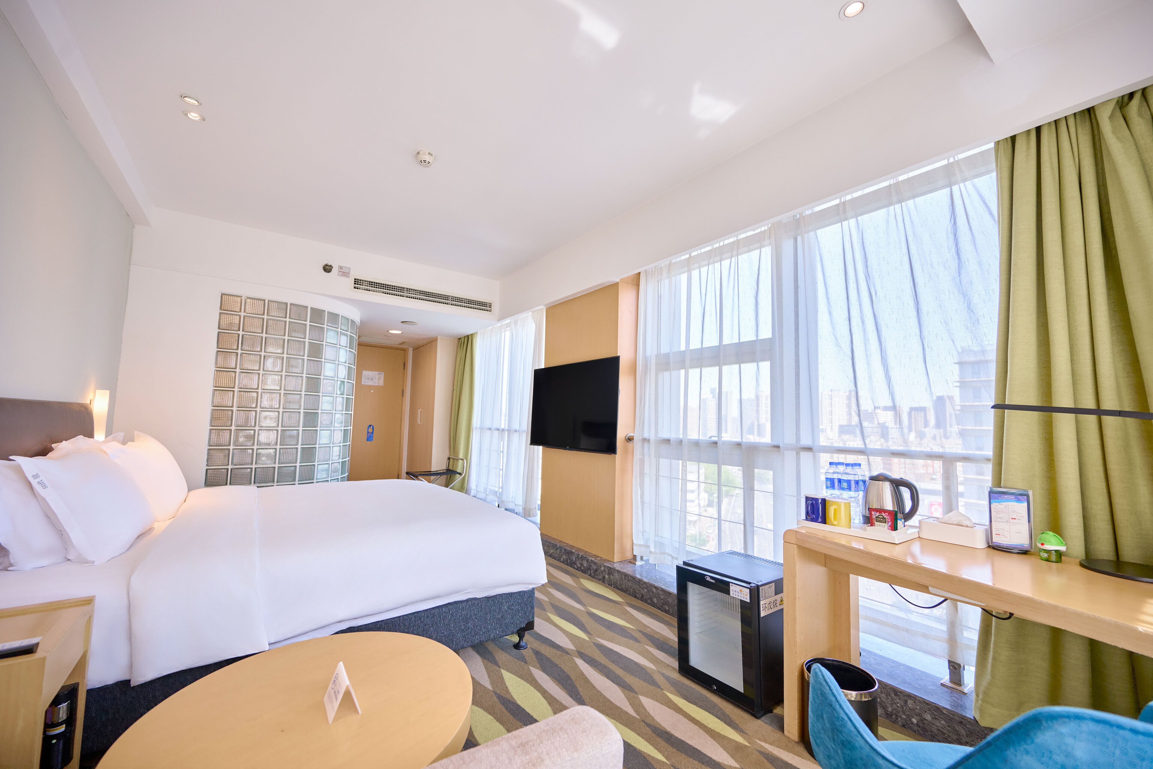 A hotel room with a bed, a desk with a computer, a small fridge, and a TV.