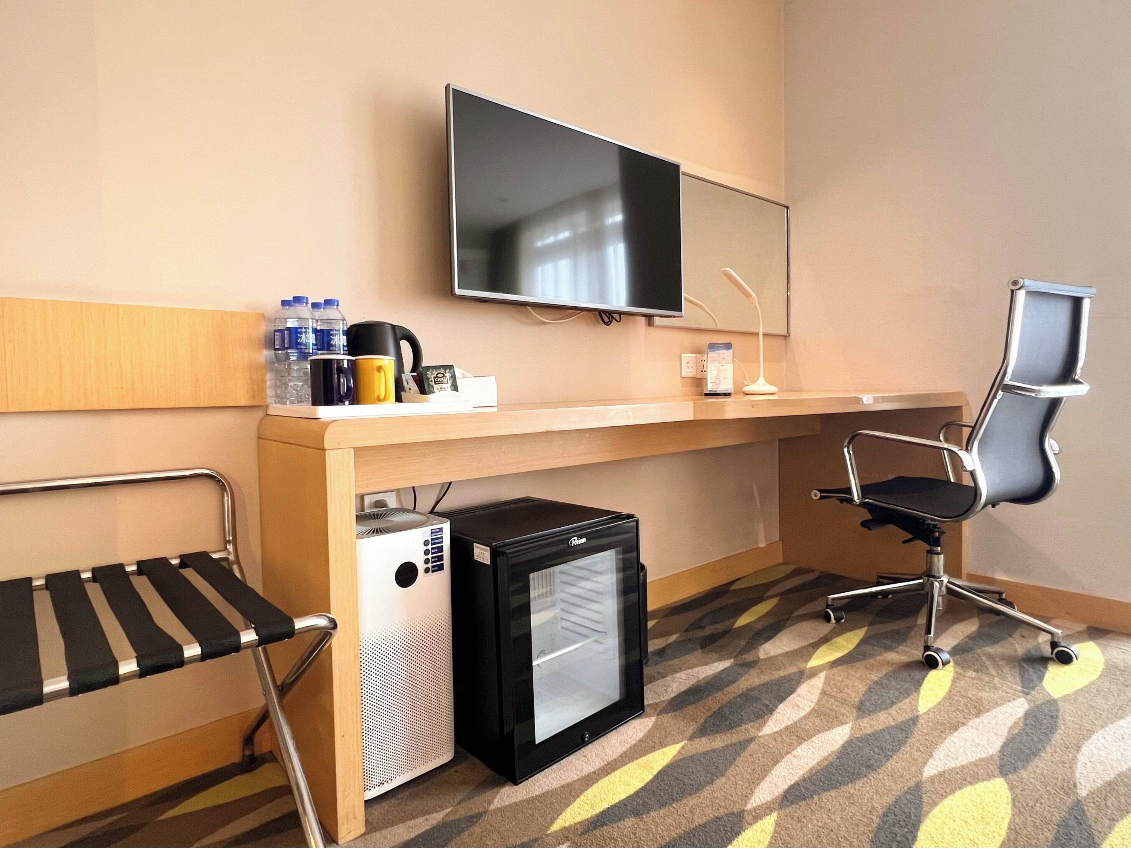 A hotel room with a desk, a chair, a television, a mini-fridge, and a bed with a striped headboard.