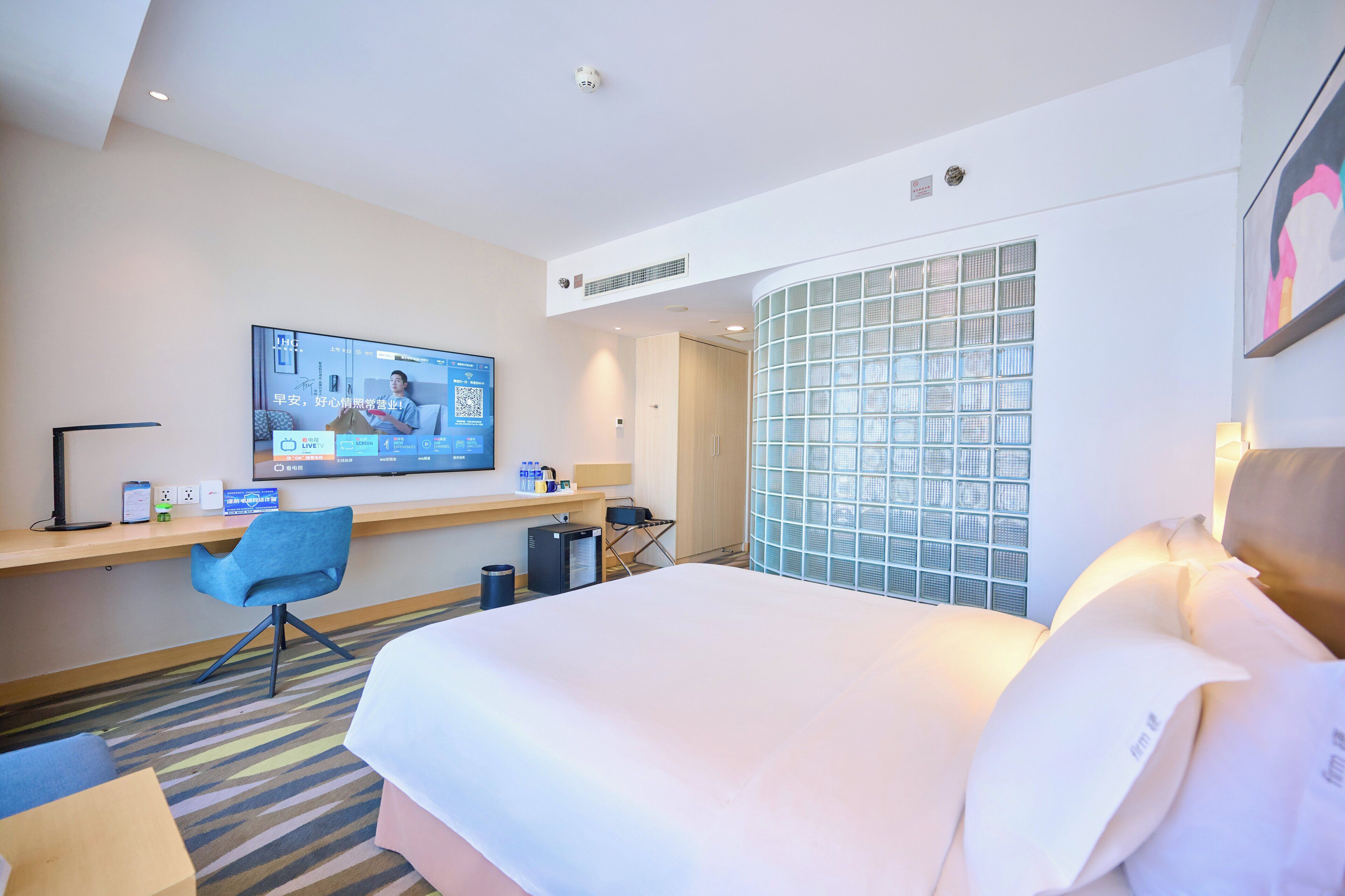 A hotel room with a large bed, a desk with a computer, a chair, and a TV displaying a live broadcast.