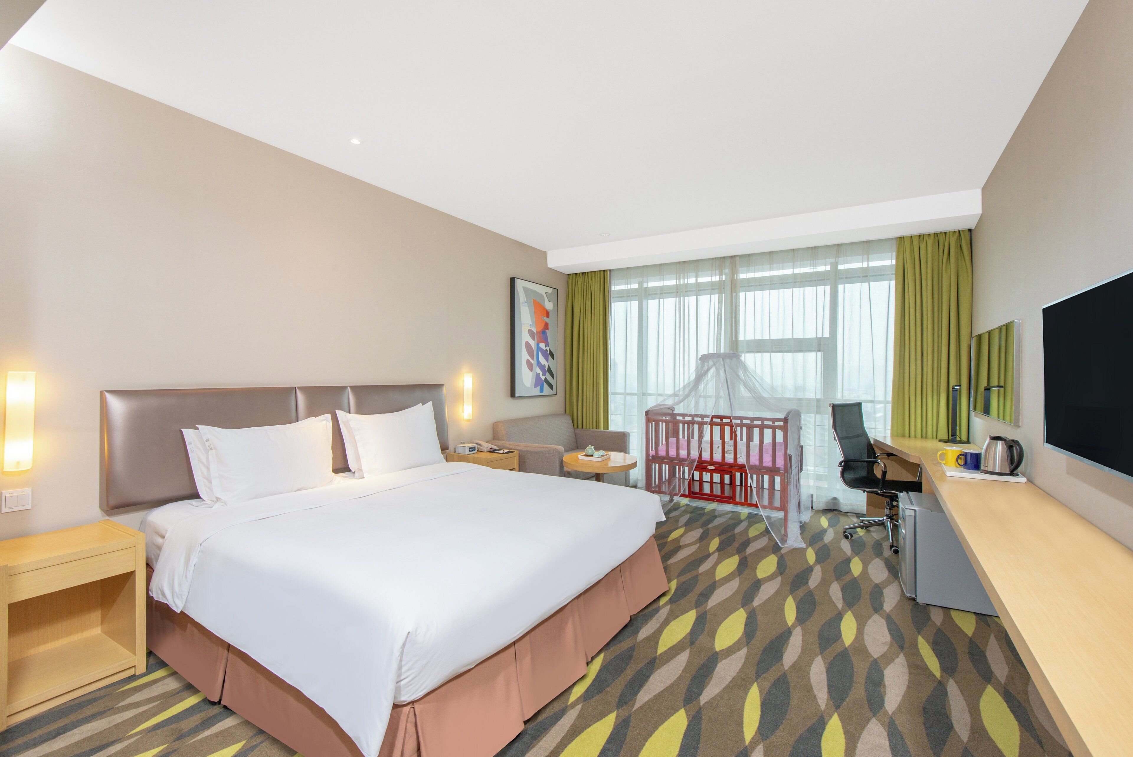 A hotel room with a large bed, bedside tables, a desk with a chair, a crib, and a large window with curtains.