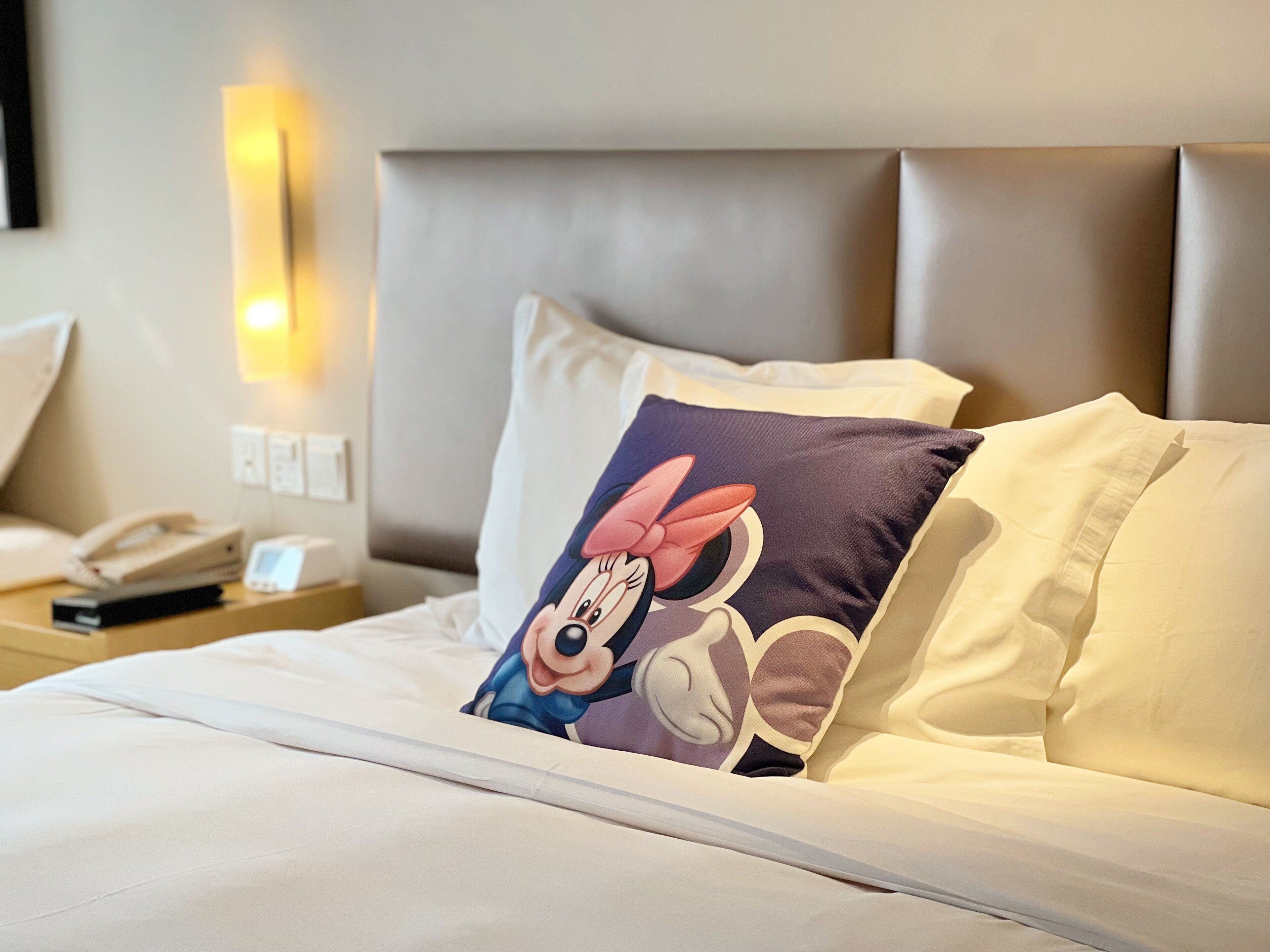 A neatly made bed with a pillow featuring a cartoon character, a bedside phone, and a wall-mounted light fixture.