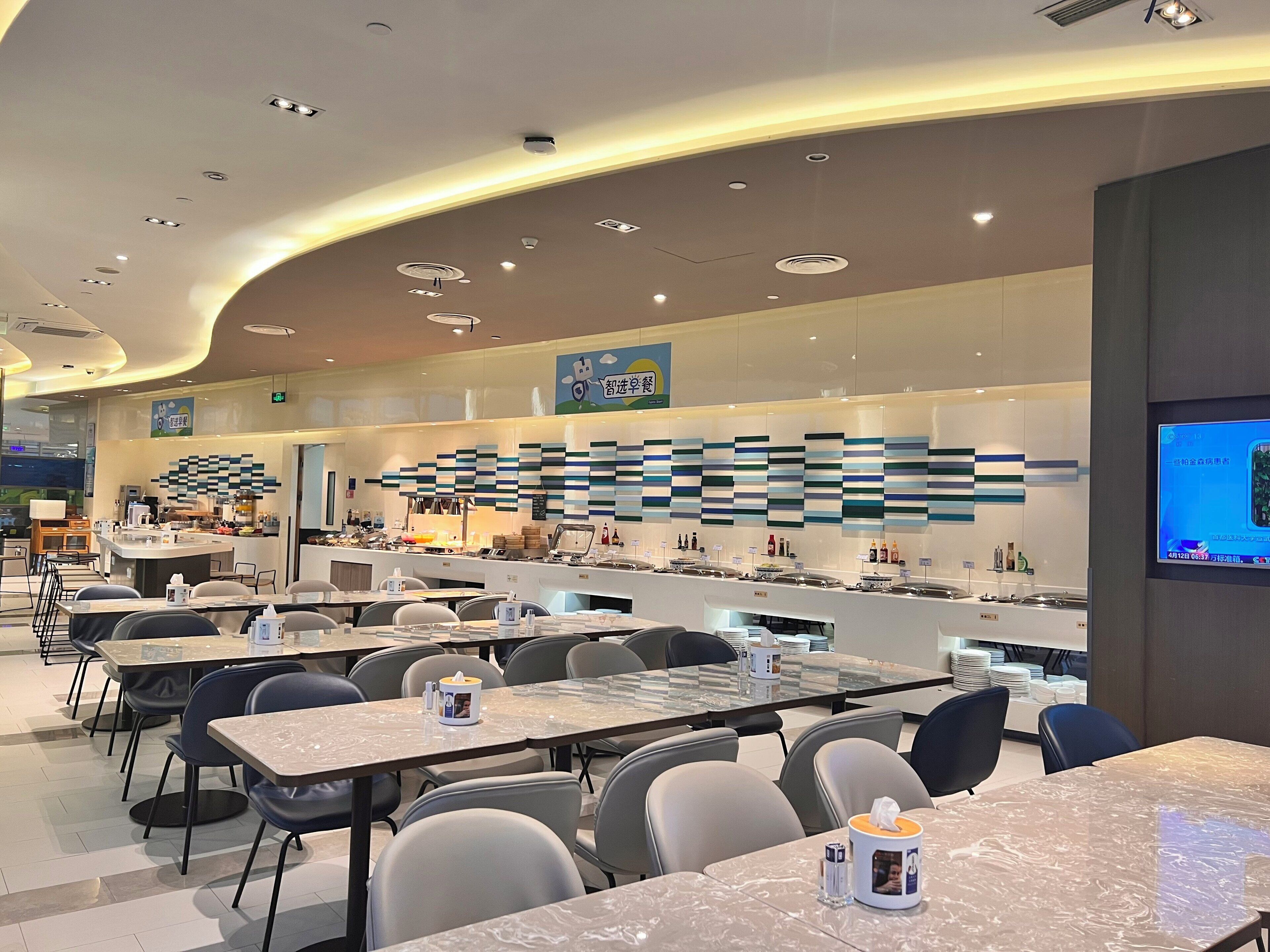 A modern restaurant with a long counter, multiple seating areas, and a digital screen displaying content.