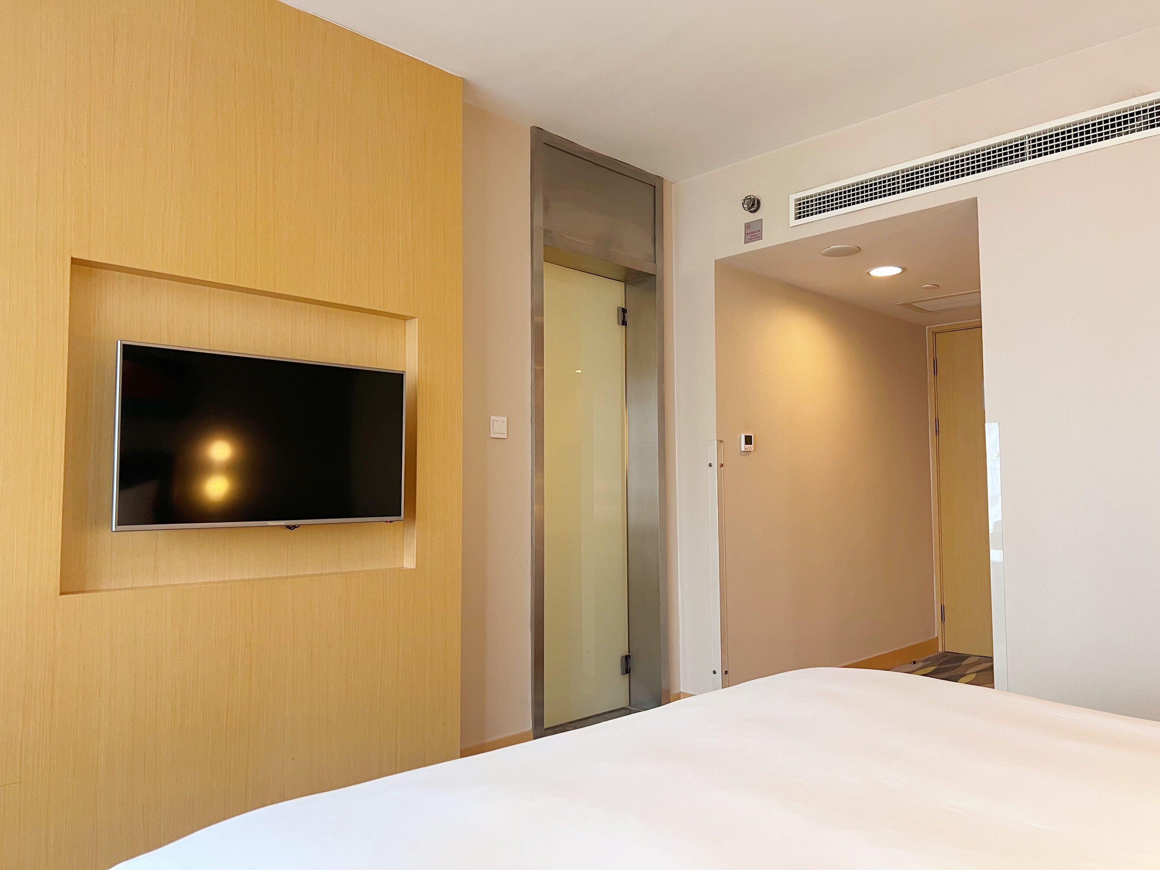 Habitación de hotel con televisión de pantalla plana montada en una pared de madera, una cama con ropa blanca y una puerta que da a otra habitación.