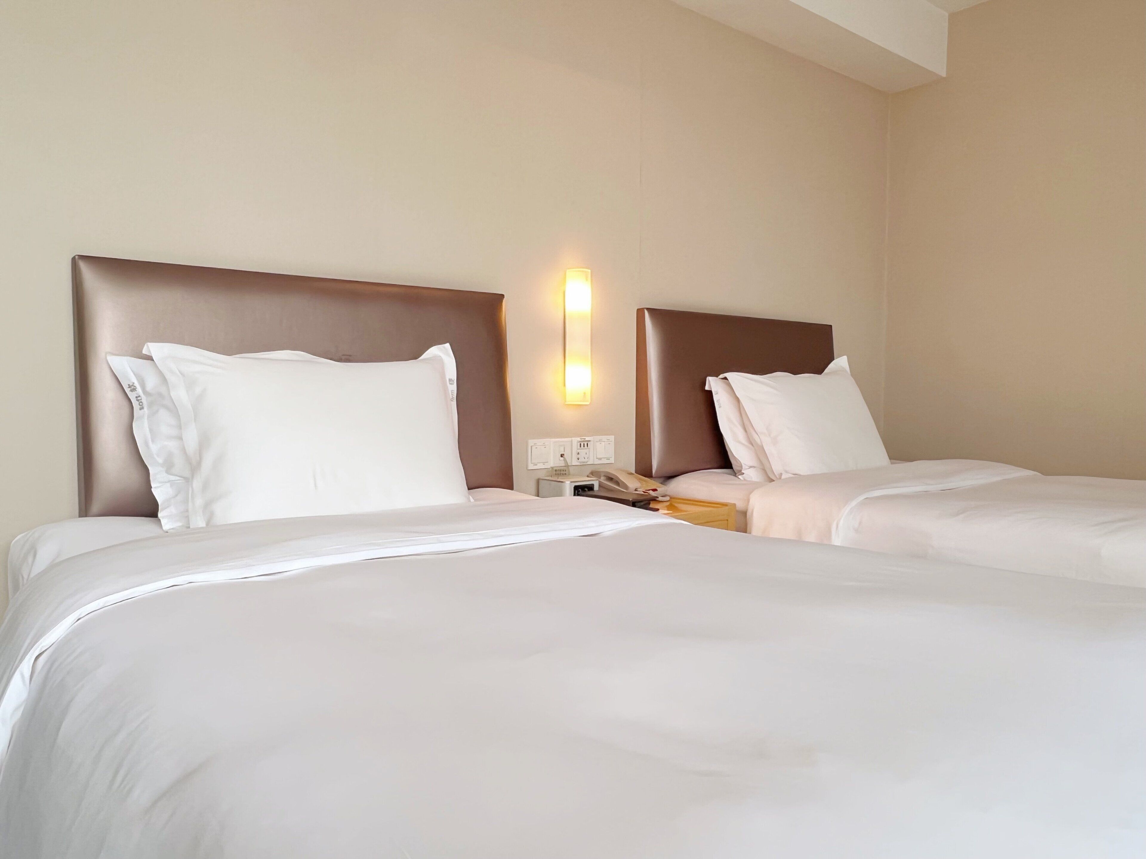 A hotel room with two beds, each with a headboard and white bedding. There is a bedside table with a lamp and a telephone.