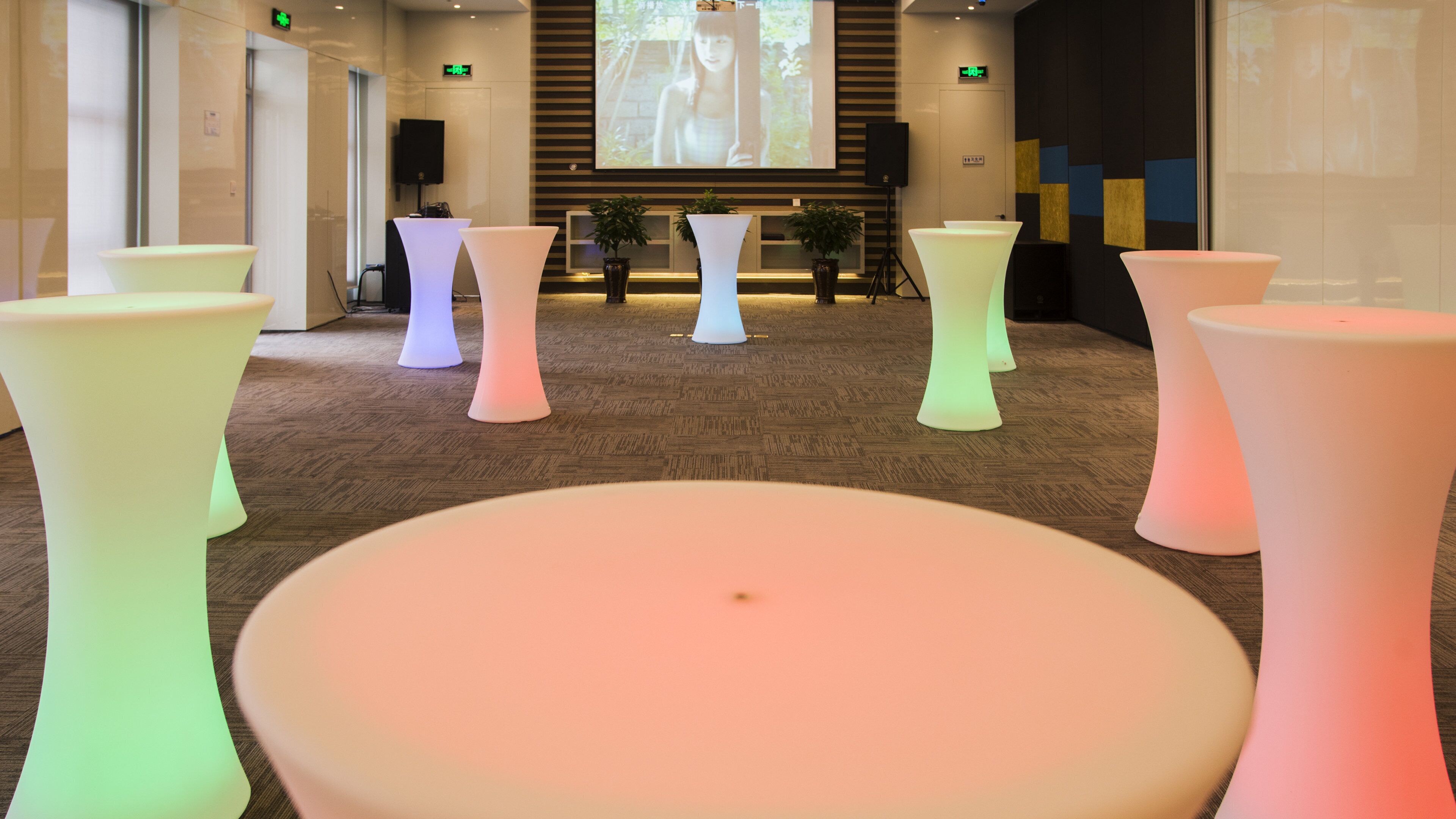 Eine moderne Lobby mit mehreren runden Tischen und Stühlen, einer Projektionsleinwand, die eine Szene zeigt, und einem Empfangsbereich mit Theke und Pflanzen.