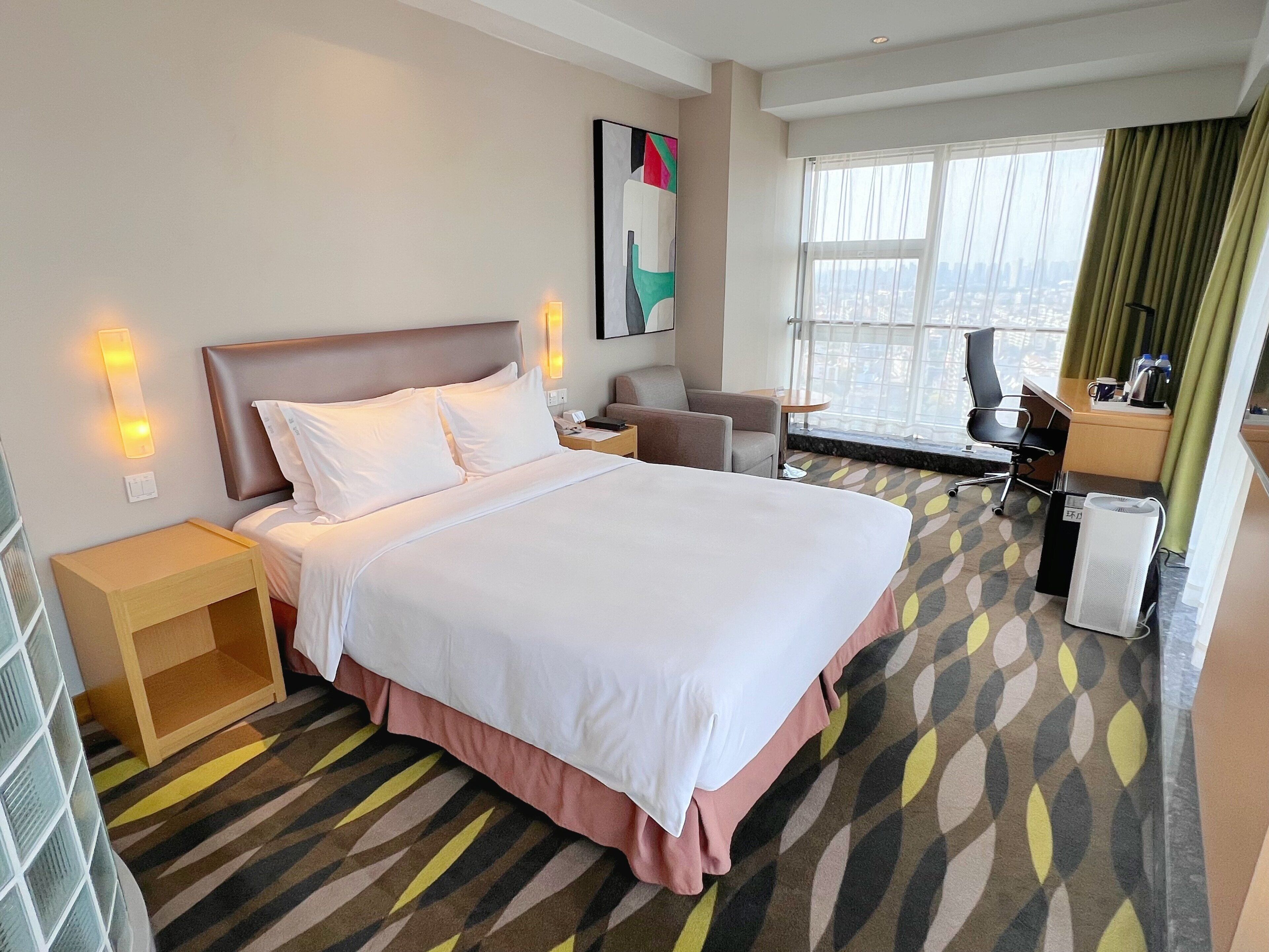 Een hotelkamer met een groot bed, een bureau, een stoel, een klein tafeltje en een raam met gordijnen.