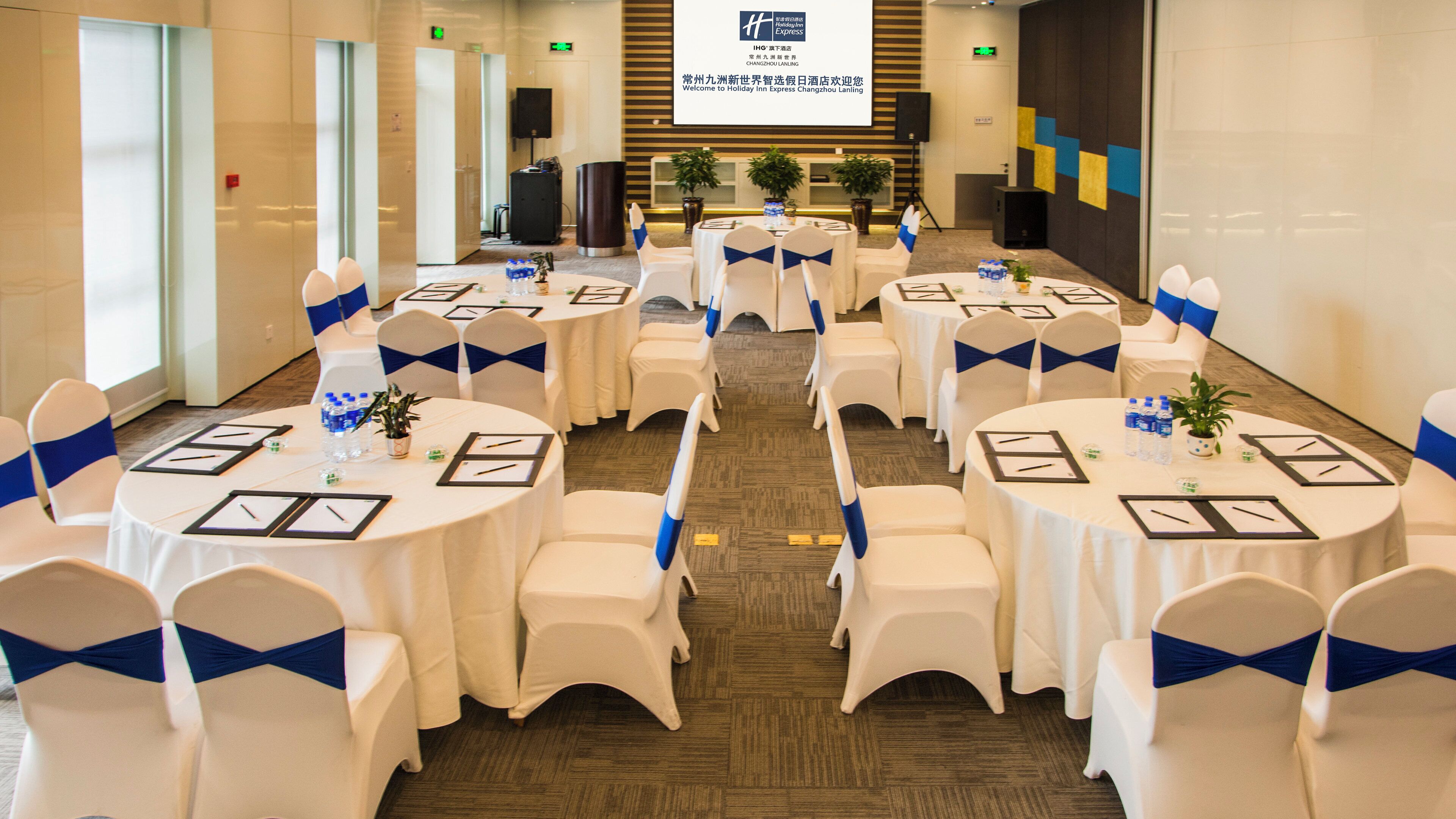 Un salón de conferencias con mesas redondas, sillas y pantallas de presentación, que sugiere una preparación para una reunión o evento.