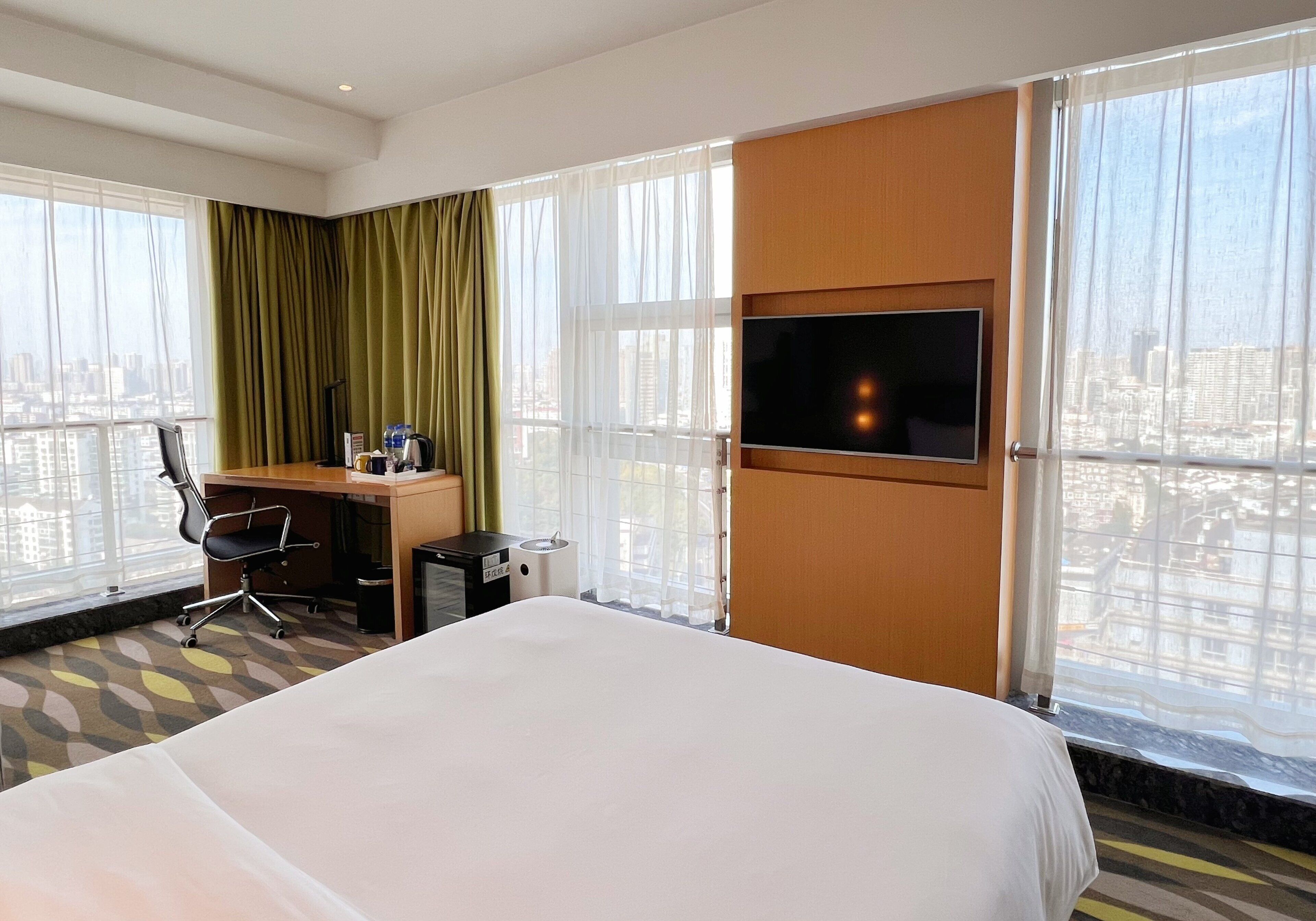 Ein Hotelzimmer mit einem großen Bett, einem Schreibtisch mit Stuhl, einem Fernseher und Blick auf die Stadt durch große Fenster.