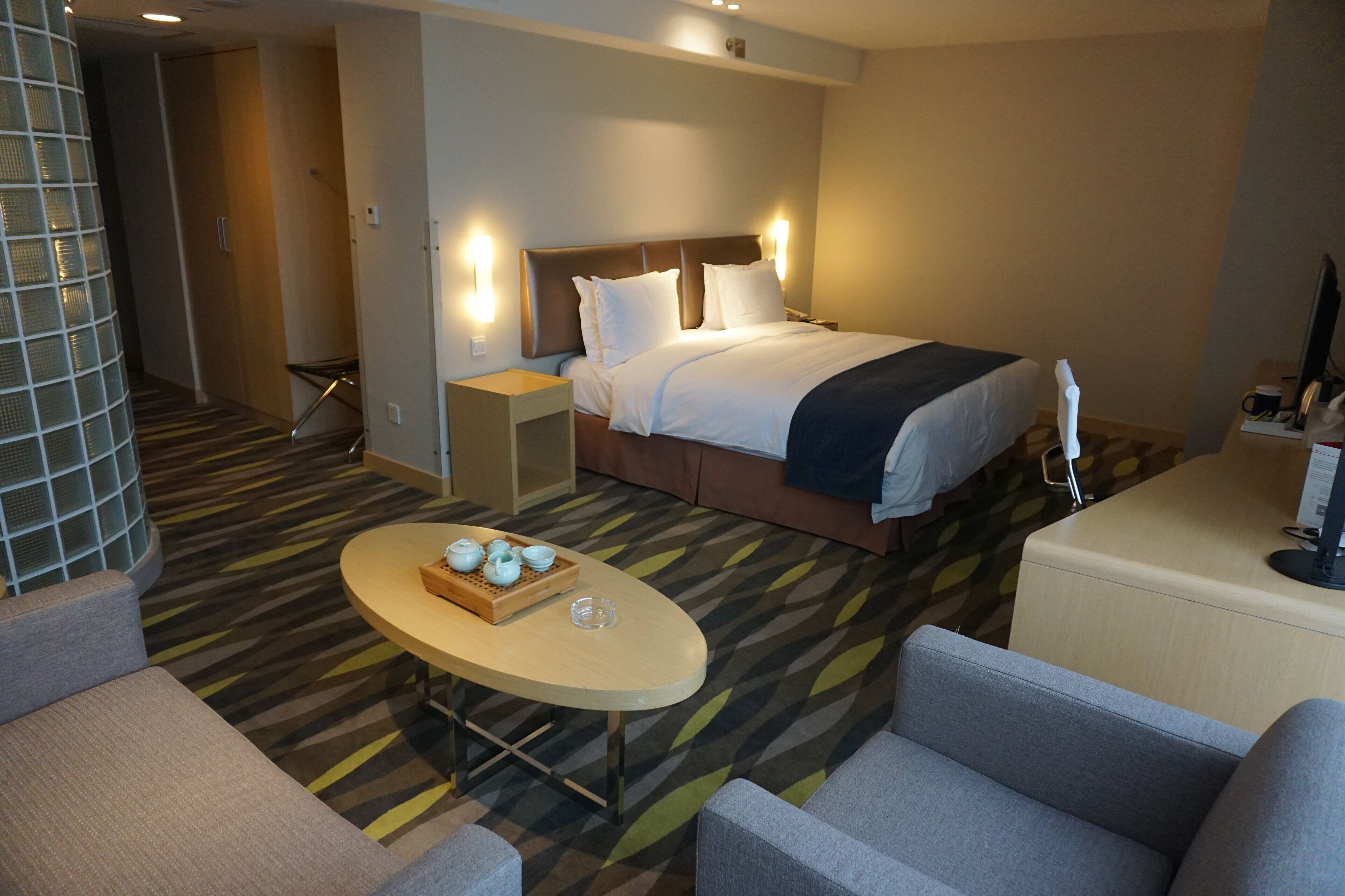 A hotel room with a bed, bedside tables, a desk, a chair, and a small table with a tray of items.