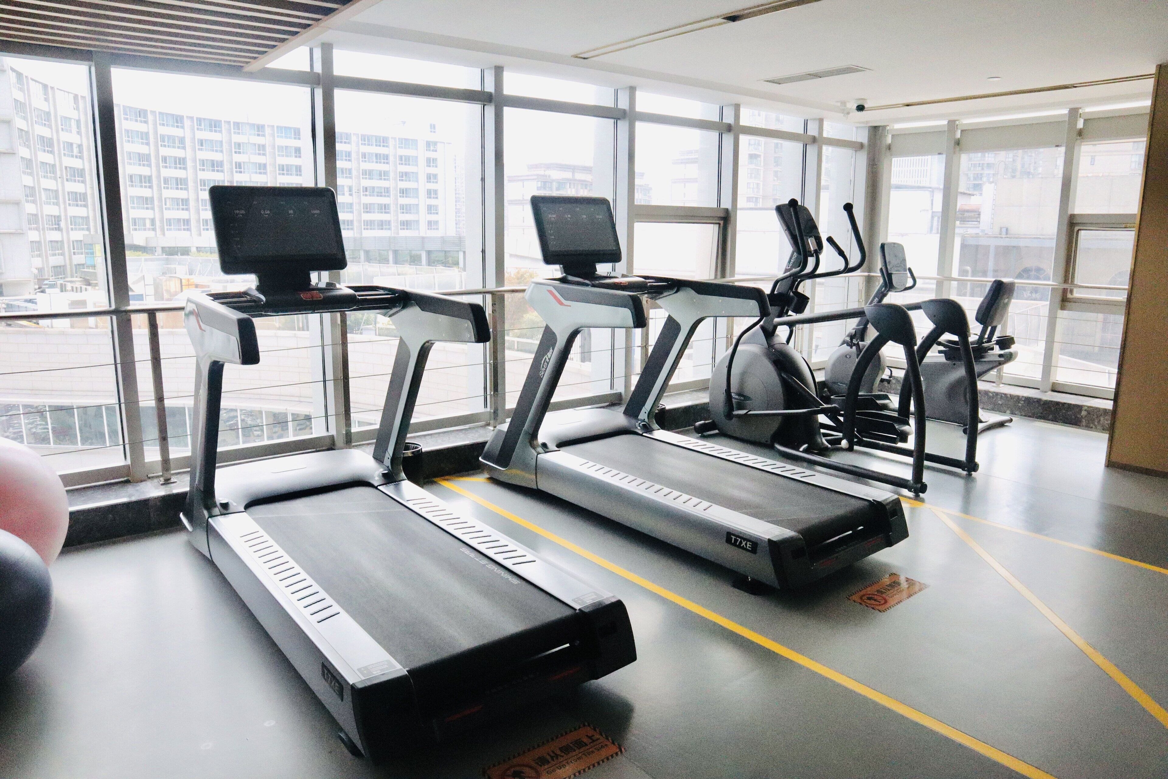 A gym with treadmills and exercise equipment, large windows, and a view of buildings outside.