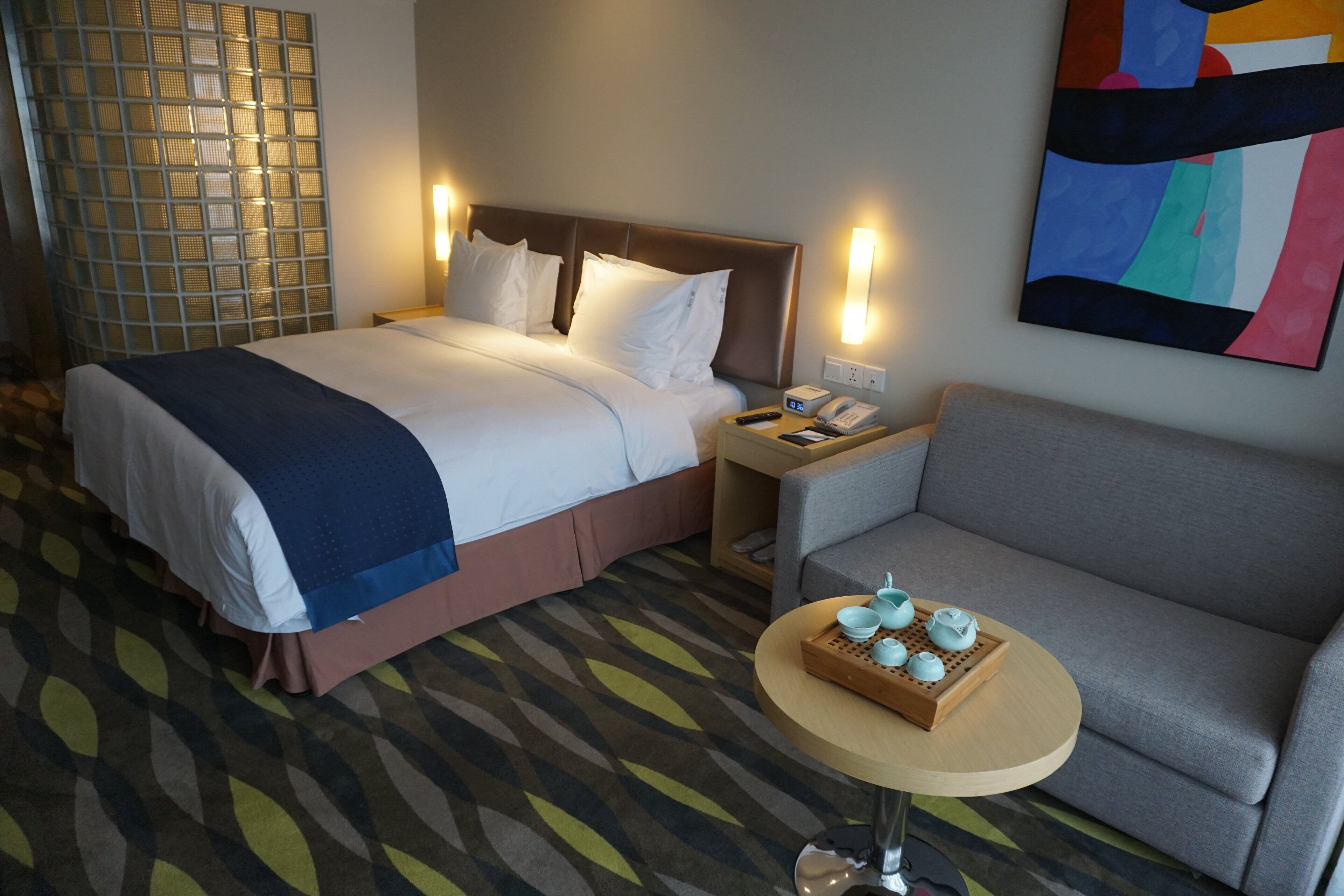 A hotel room with a bed, bedside table, a small table with teapots, and a grey sofa.