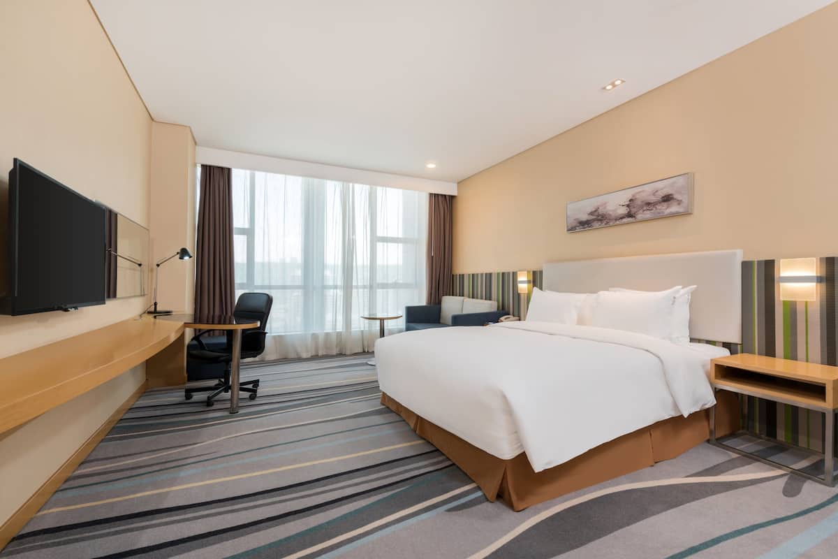 Une chambre d’hôtel avec un grand lit, un bureau, une chaise, une télévision et une fenêtre avec des rideaux.