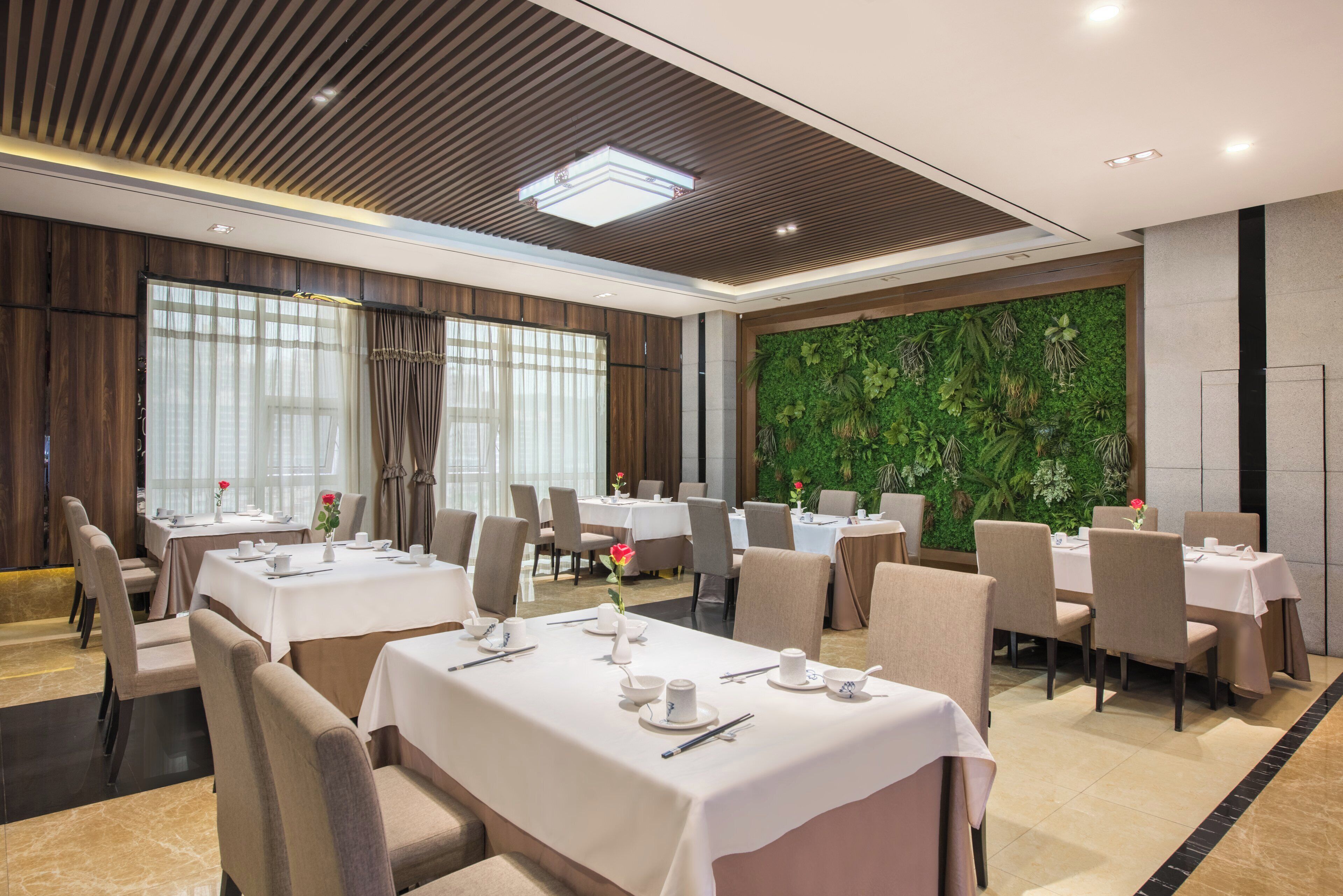 Un comedor de hotel con mesas dispuestas para el servicio, un ventanal con cortinas y una pared verde con plantas.