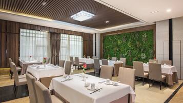Un comedor de hotel con mesas dispuestas para el servicio, un ventanal con cortinas y una pared verde con plantas.