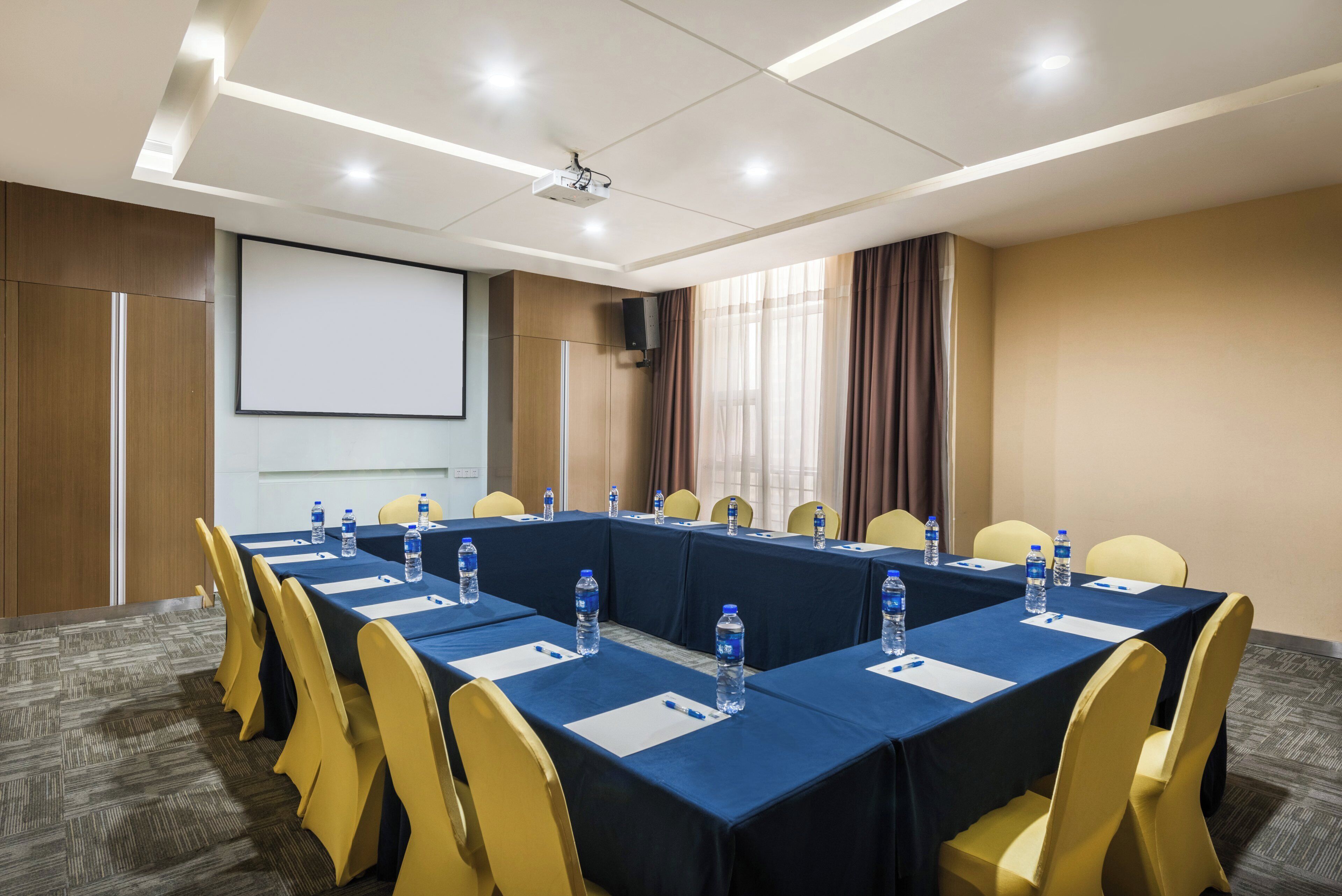 Un salón de conferencias con una larga mesa, sillas amarillas, botellas de agua y una pantalla de proyector.