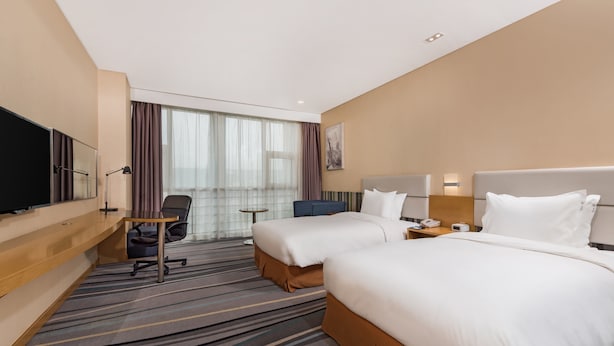 Une chambre d’hôtel avec deux lits, un bureau, une chaise, une télévision et une grande fenêtre avec des rideaux.