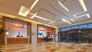 Uma área de recepção de hotel com design moderno, incluindo um balcão de atendimento, um painel digital e uma entrada de vidro.