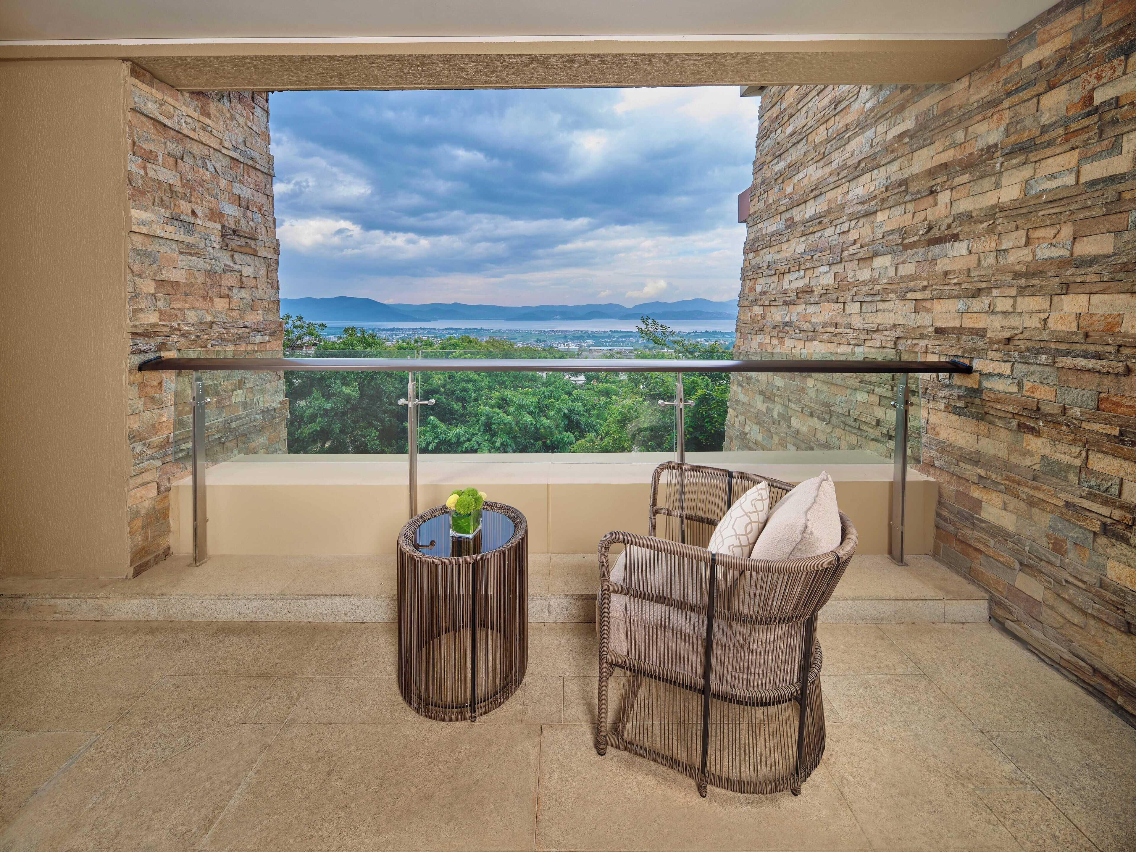 Ein Balkon mit Steinmauer, einem Korbstuhl, einem kleinen Tisch mit einer Vase und Blick auf Grünflächen und Berge.