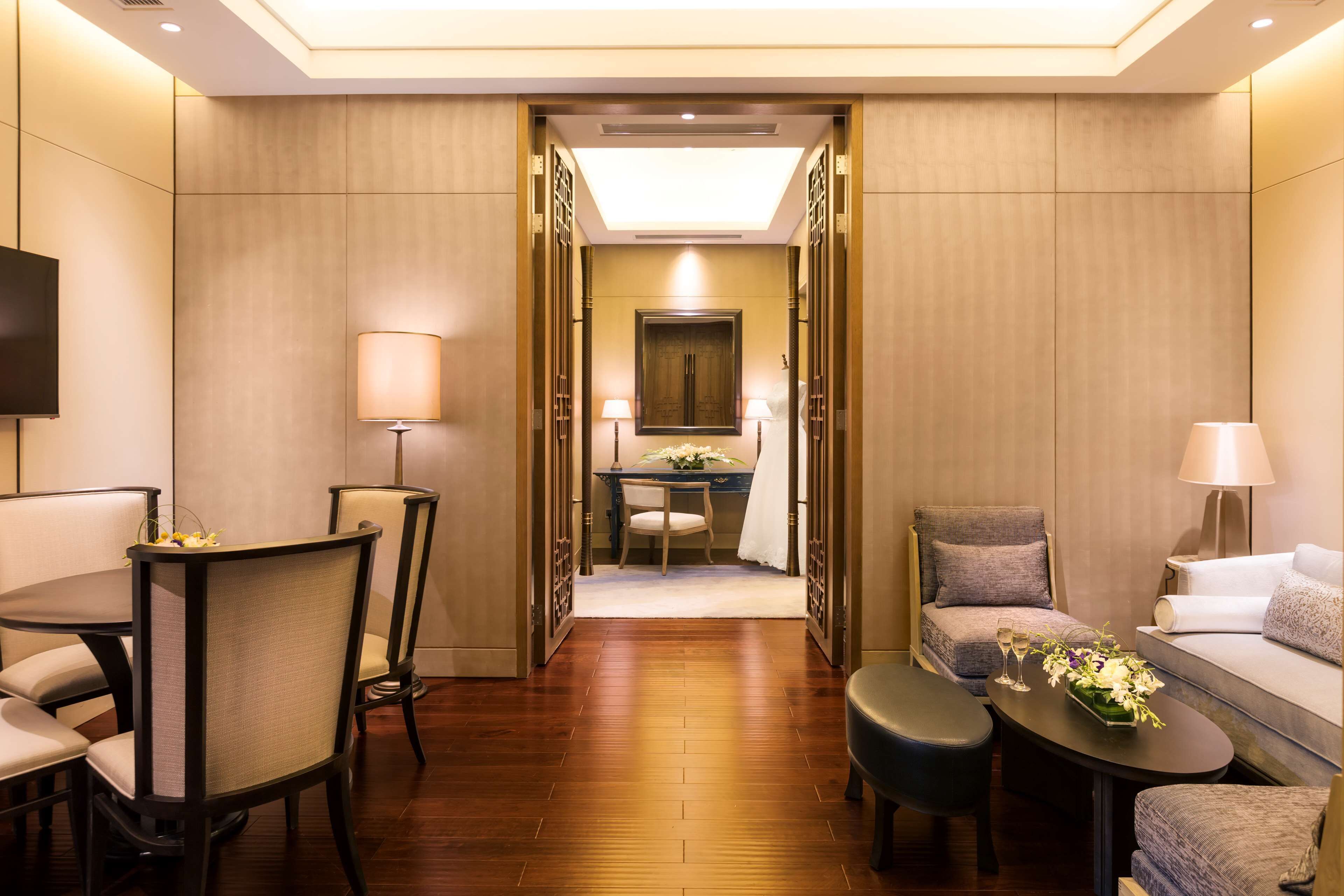 A hotel room with a wooden floor, a dining area with a table and chairs, a sofa, a TV, and a desk with a lamp.