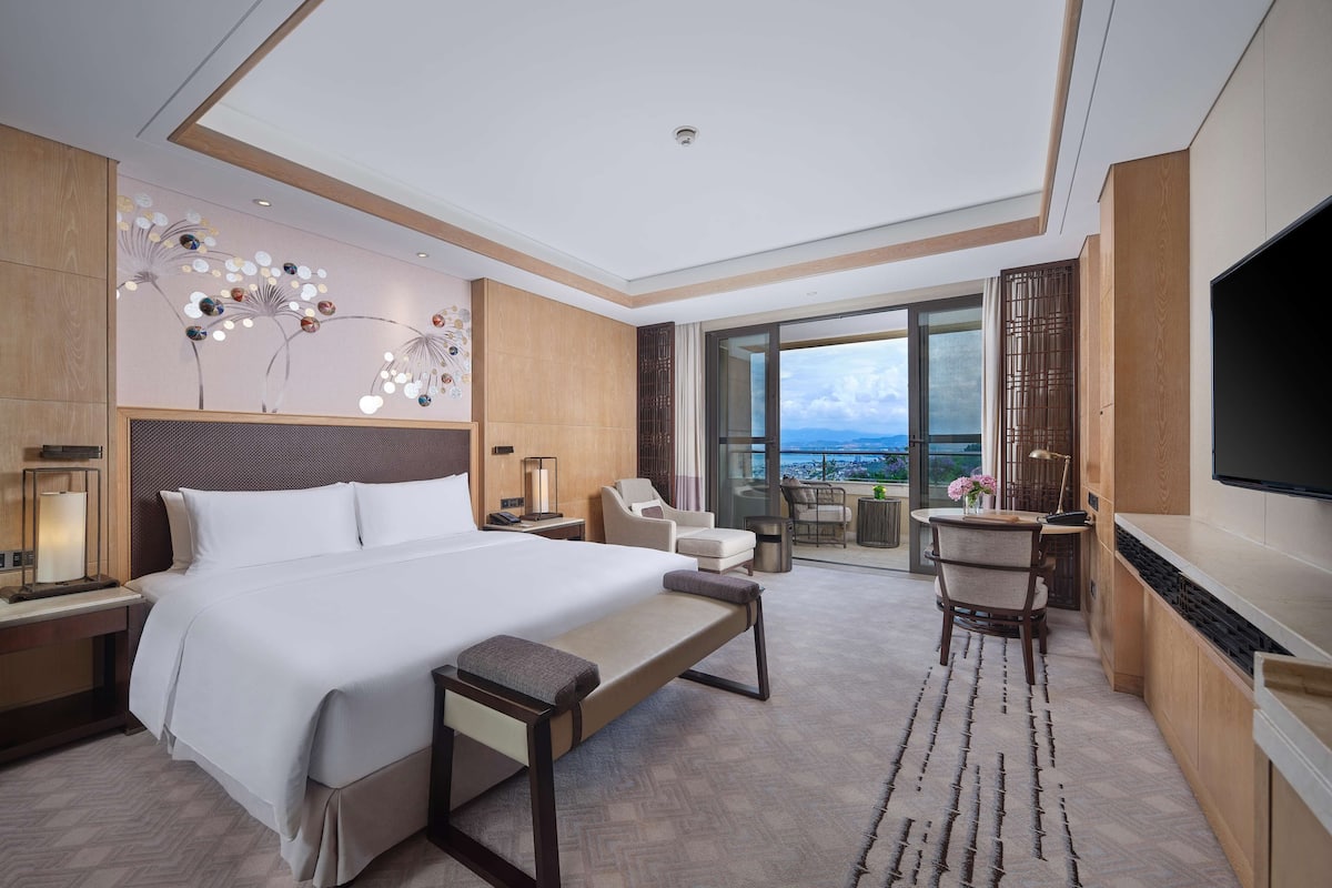 Quarto de hotel moderno com uma cama grande, uma escrivaninha, uma cadeira, televisão e varanda com vista para a cidade.