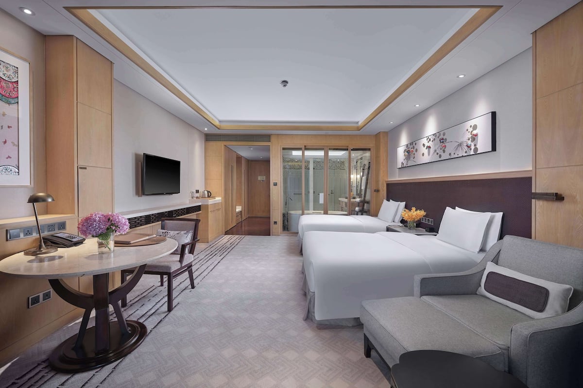 Quarto de hotel com cama, sofá, escrivaninha com luminária, televisão e banheiro visível por uma porta de vidro.