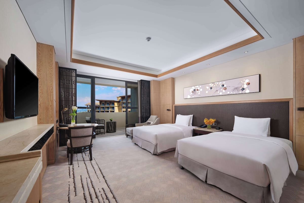 Quarto de hotel com duas camas, uma escrivaninha, televisão e varanda com vista para outros edifícios.