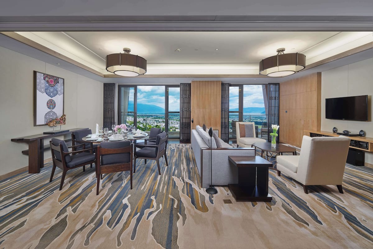 Quarto de hotel com área de refeições, sofá, televisão e uma grande janela com vista para a cidade.