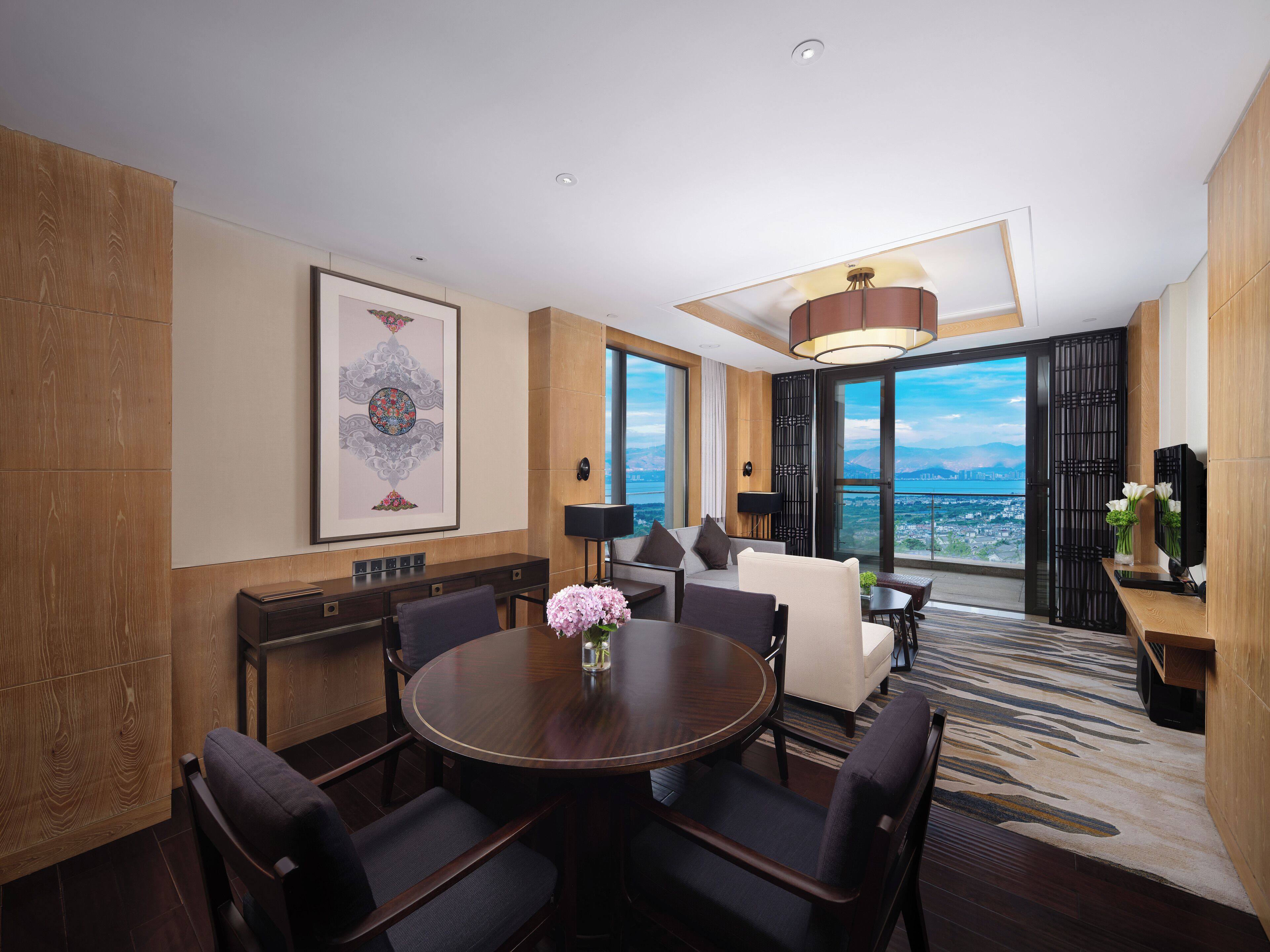Una habitación de hotel moderna con comedor, sofá, ventana grande con vista a la ciudad y un adorno de pared decorativo.