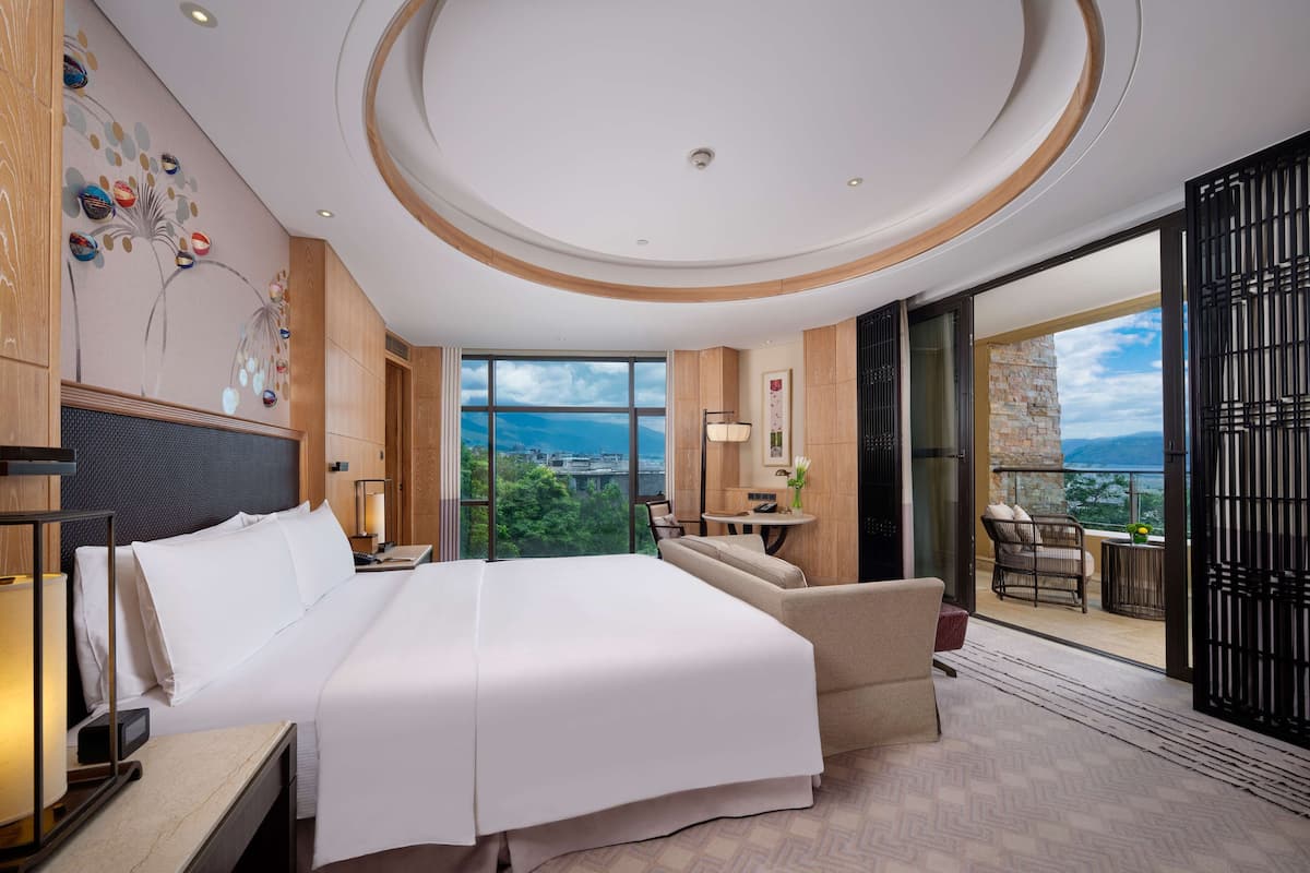 Quarto de hotel moderno com uma cama grande, um espaço de estar, varanda com vista e paisagem urbana visível pelas janelas.