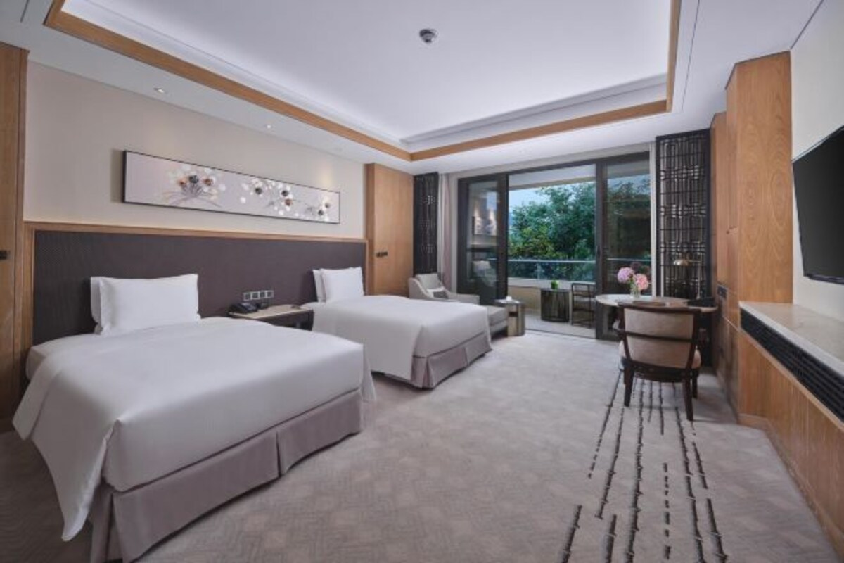 A hotel room with two beds, a desk, a chair, a TV, and a balcony with a view.