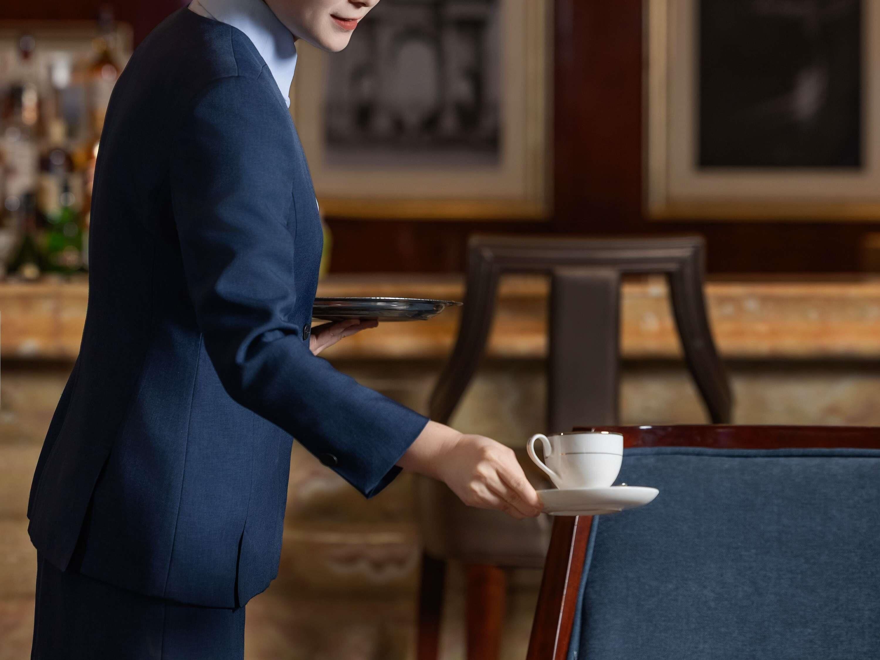 Eine Person in einem blauen Anzug serviert eine weiße Tasse und Untertasse auf einem Tablett.