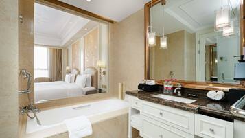 Een hotelbadkamer met een grote spiegel, een bad, een wastafelmeubel met een wastafel, en een bed dat door de spiegel zichtbaar is.