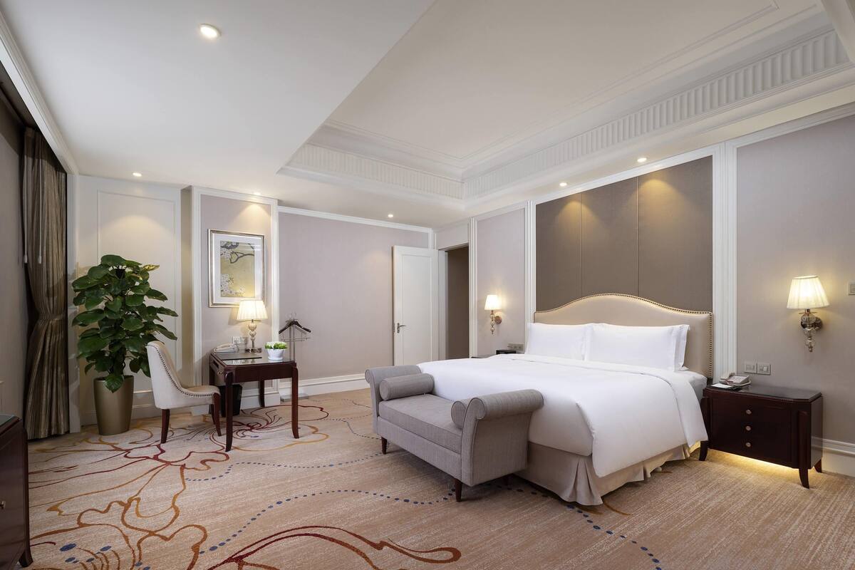 A hotel room with a large bed, a desk with a lamp, a chair, a potted plant, and a nightstand with a lamp.