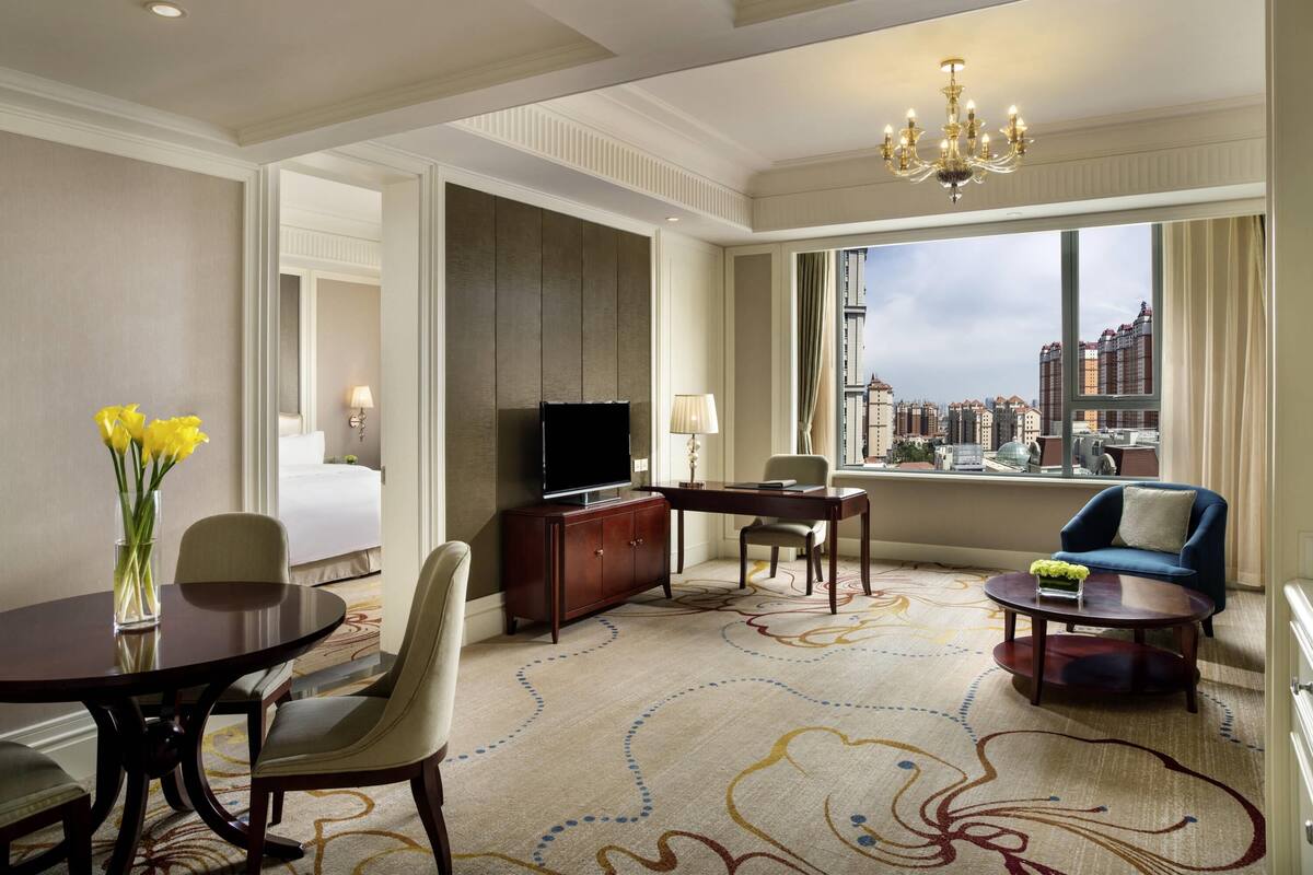 A hotel room with a bed, a TV, a dining table, a sofa, a desk, and a view of the city.