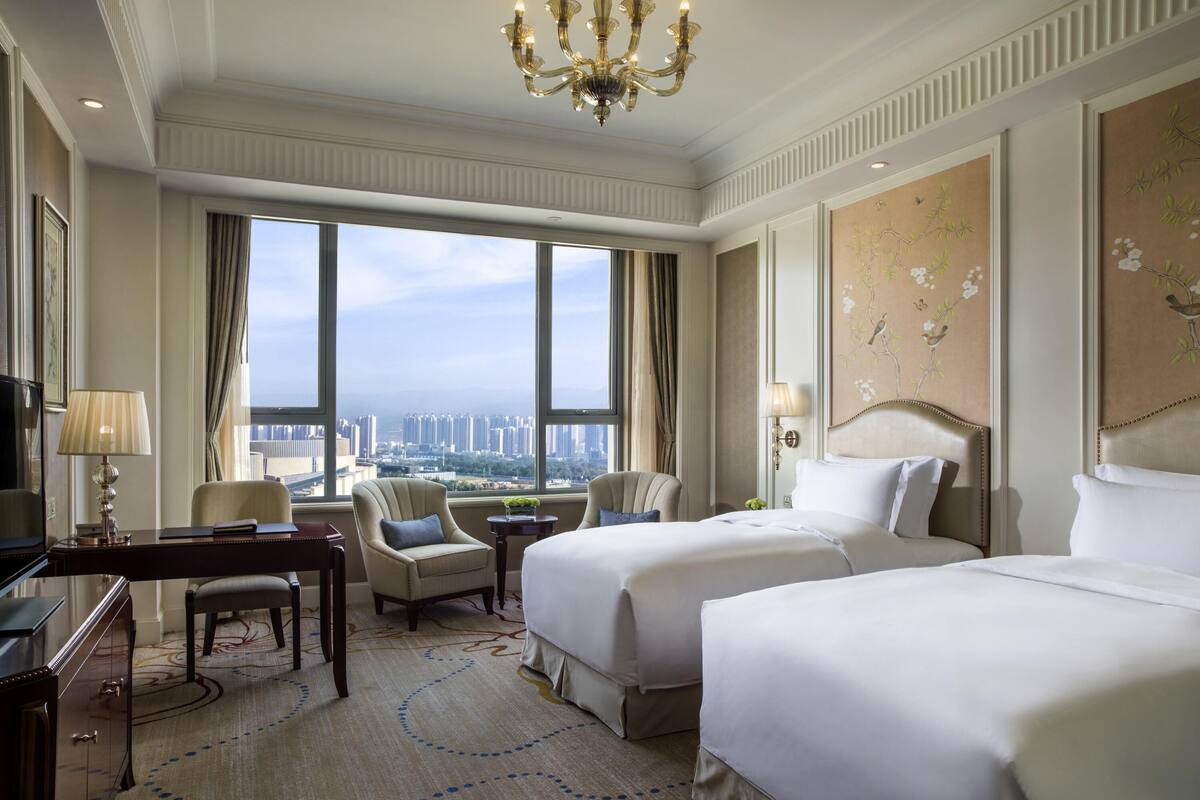 A hotel room with two beds, a desk, a chair, a chandelier, and a large window with a city view.