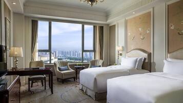 A hotel room with two beds, a desk, a chair, a chandelier, and a large window with a city view.