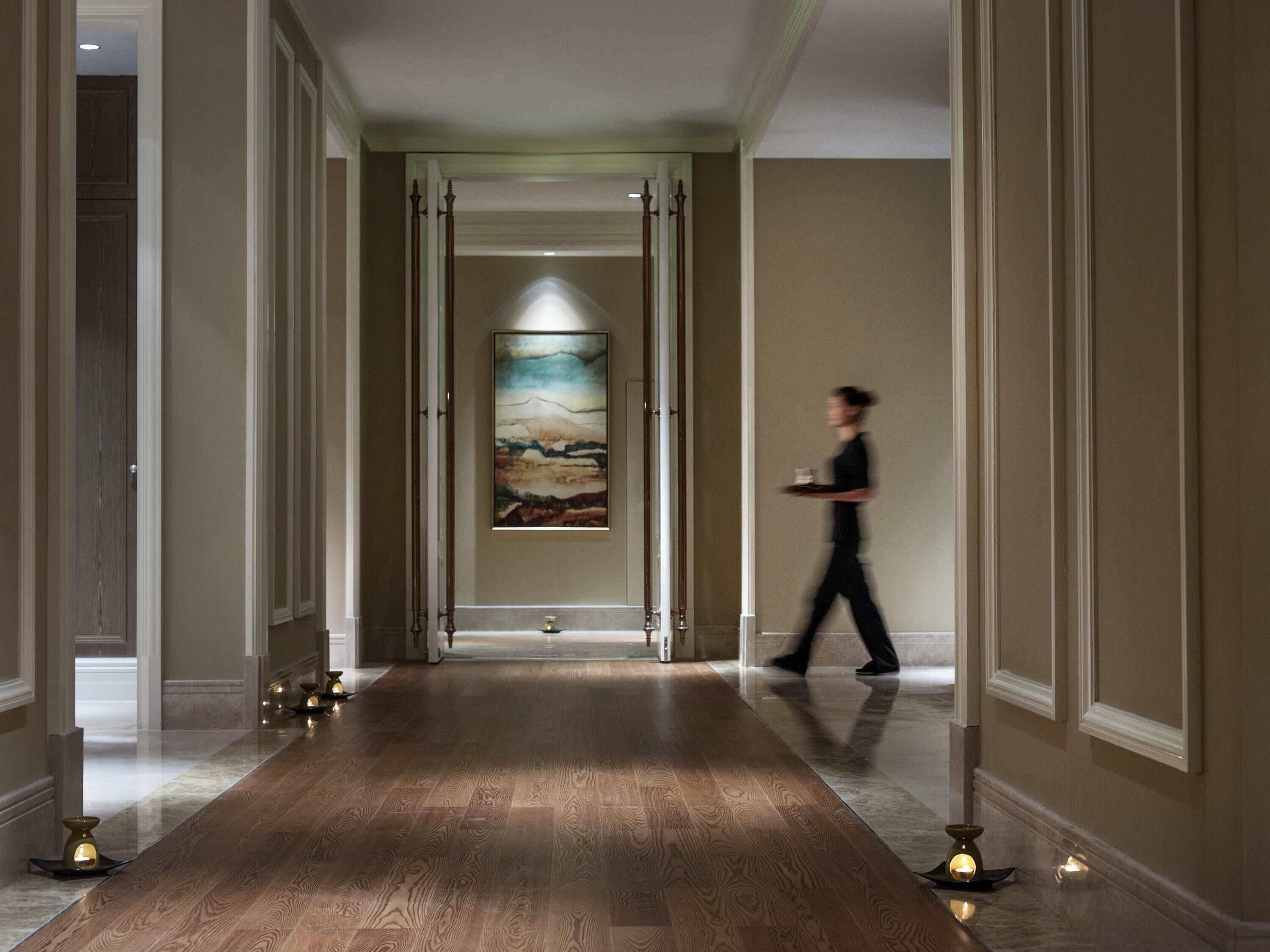 A hotel corridor with a wooden floor, a painting on the wall, and a person walking.