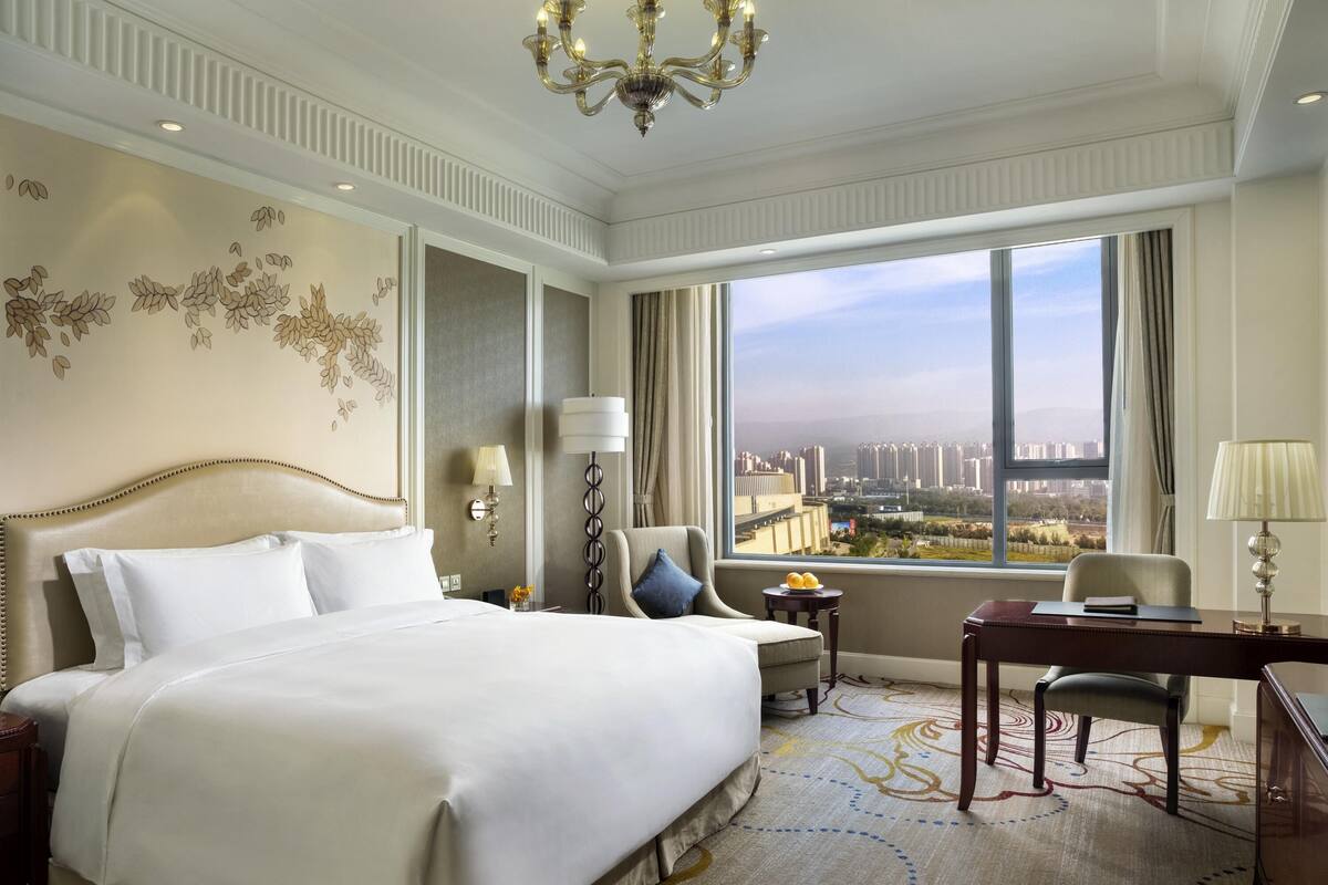 A hotel room with a large bed, a desk, a chair, a lamp, a side table, a wall-mounted lamp, and a view of a cityscape through large windows.