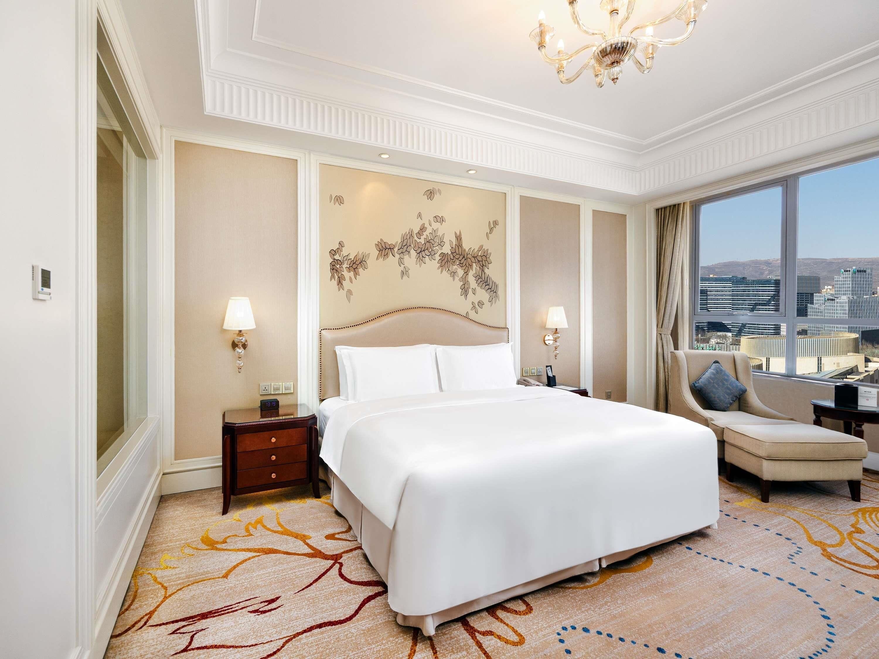 A hotel room with a large bed, a sofa, a view of the city, and a chandelier.