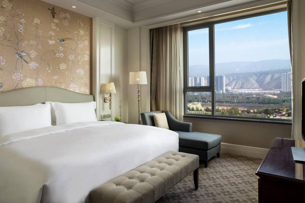 A hotel room with a large bed, a desk, a chair, a lamp, and a view of a cityscape through a large window.