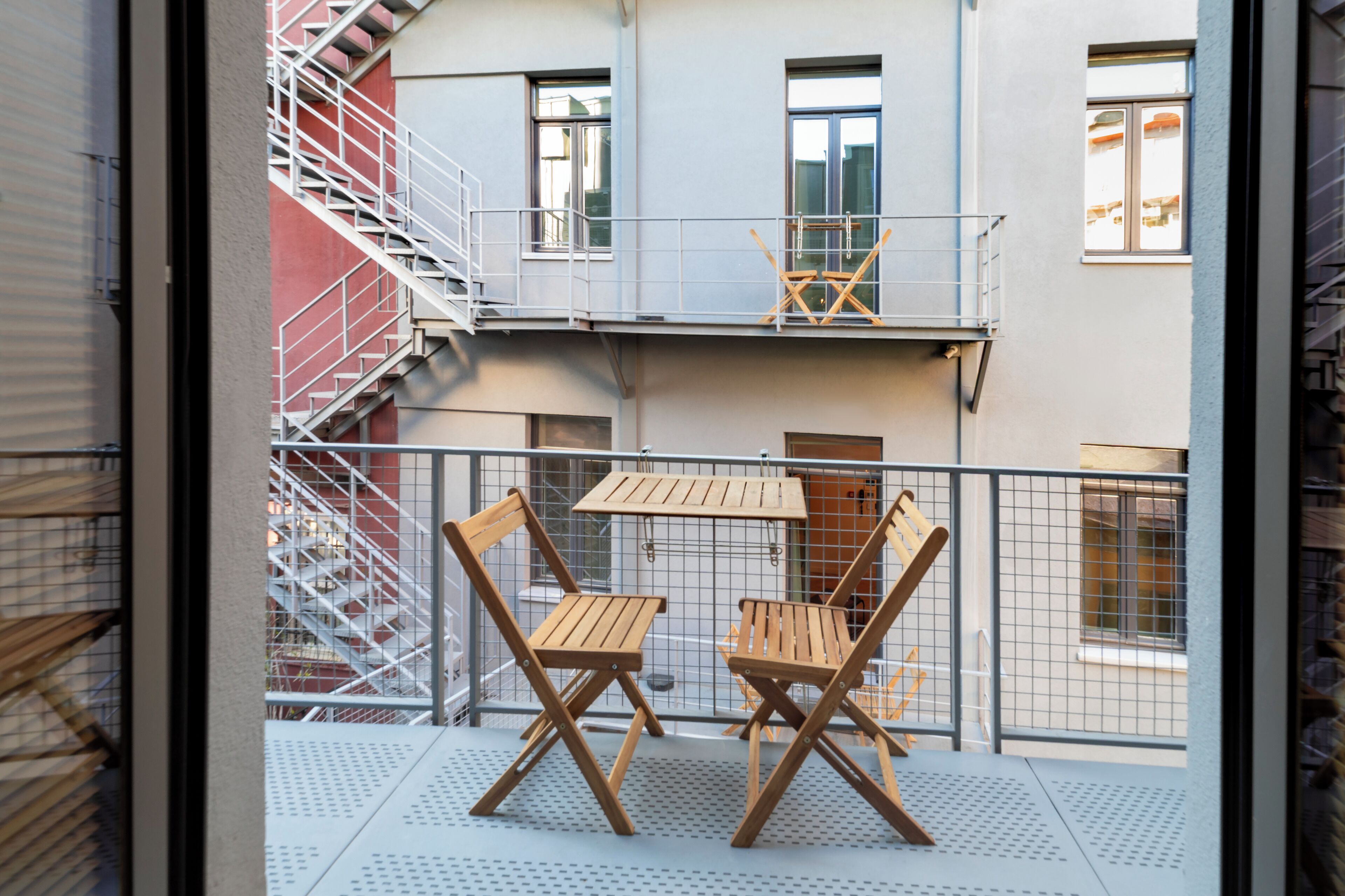 Un balcone con due sedie di legno e un tavolo, una ringhiera in metallo e una scala antincendio a sinistra.