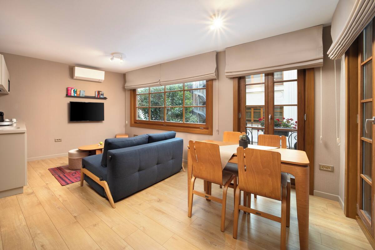 Un salon moderne avec un canapé, une table à manger et des chaises. Un téléviseur est fixé au mur, il y a une fenêtre donnant sur un espace verdoyant, et un petit coin cuisine avec un grille-pain.