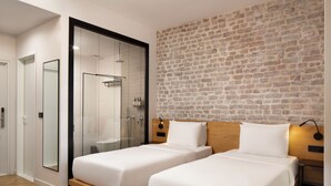 Une chambre d’hôtel moderne avec deux lits simples, un grand miroir, une douche en verre et un mur décoré d’un motif en briques.