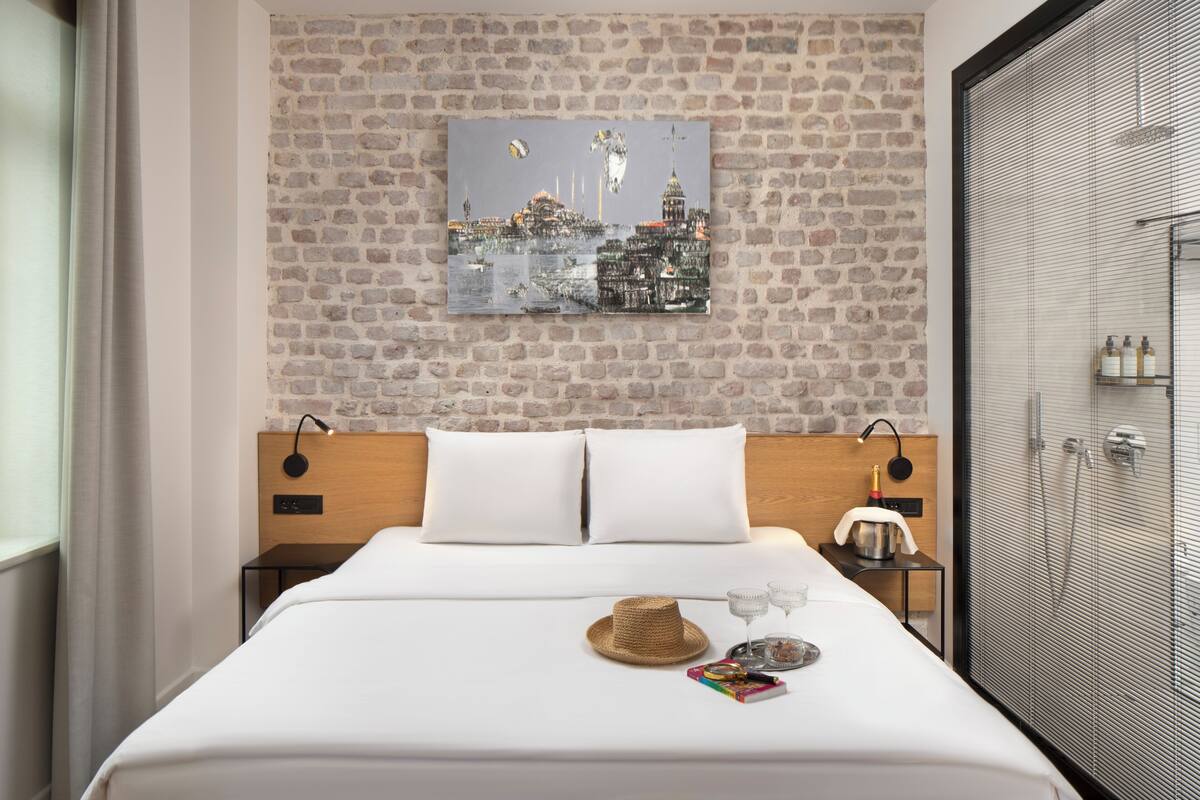 Une chambre d’hôtel avec un mur en briques, un lit avec du linge de maison blanc, une tête de lit en bois, une table de chevet avec une lampe, une cabine de douche en verre et un tableau représentant un paysage urbain accroché au mur.